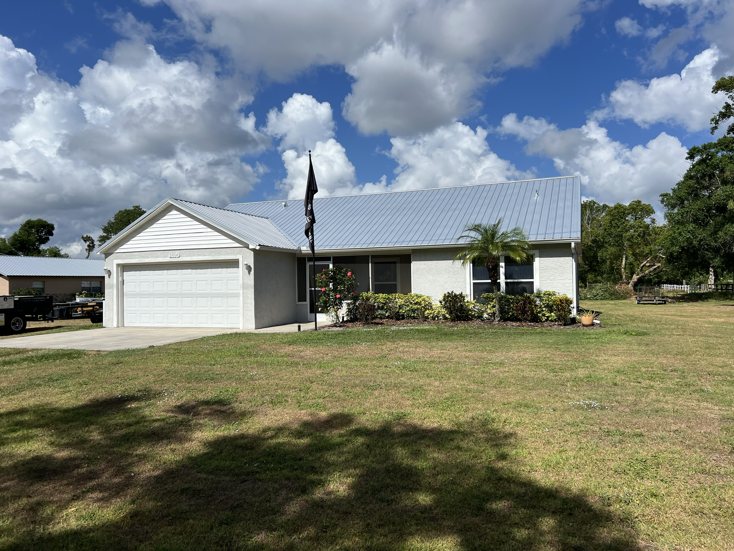 Single-story white house with a metal roof, front yard with grass, small palm tree, bushes, a black flag, and a driveway leading to a two-car garage on a partly cloudy day.
