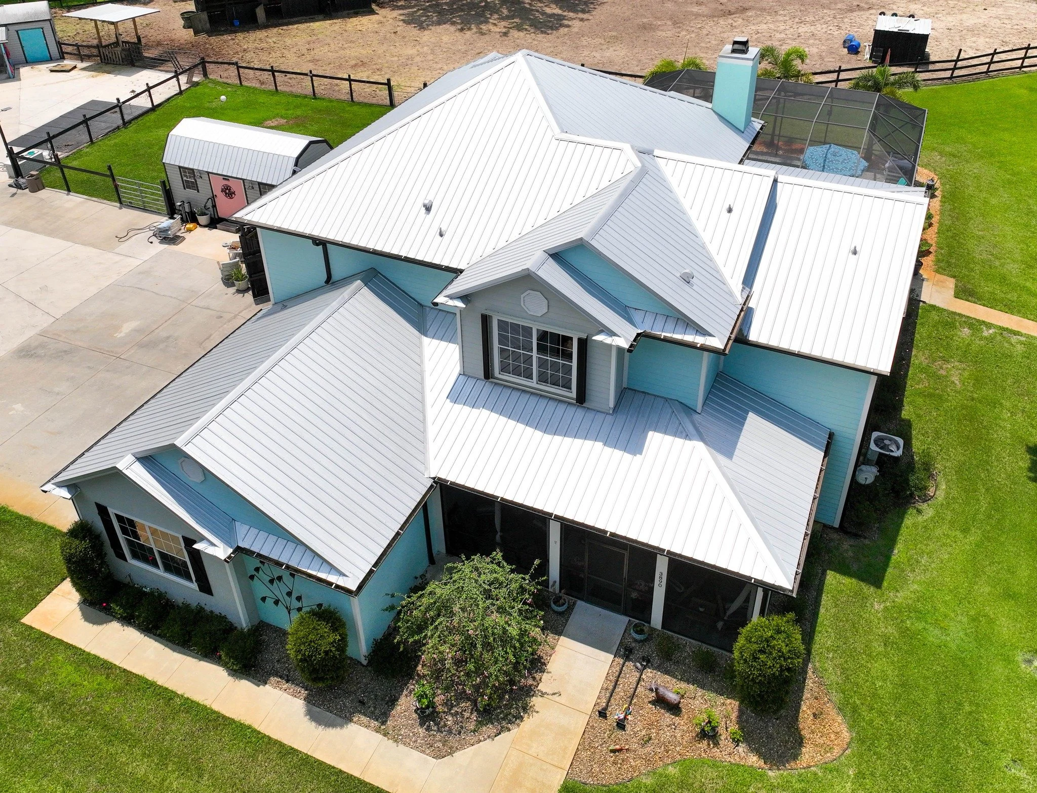 Aerial view of a house with a white metal roof, blue walls, surrounded by a lawn, concrete walkways, and a fenced backyard with a pool enclosure.