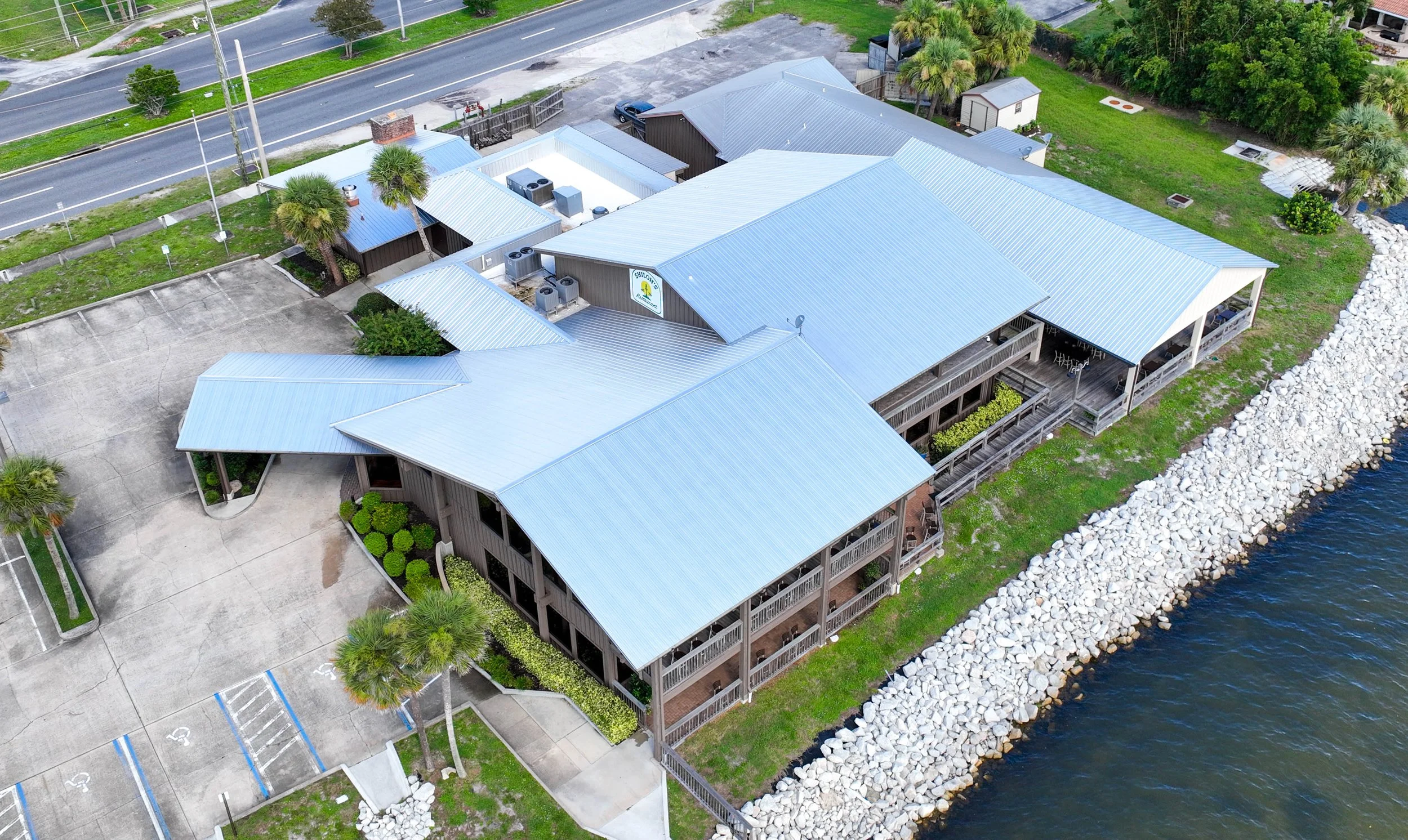 Aerial view of a large waterfront building with multiple connected sections, metal roofs, a parking lot, landscaped areas with palm trees, and a rocky shoreline along a body of water.