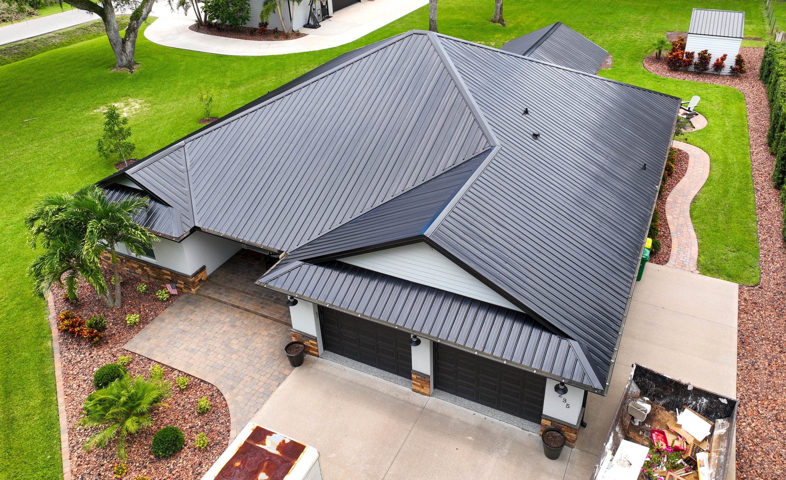 An aerial view of a modern house with a dark metal roof, a driveway, landscaped yard with green grass, trees, shrubs, and garden beds.
