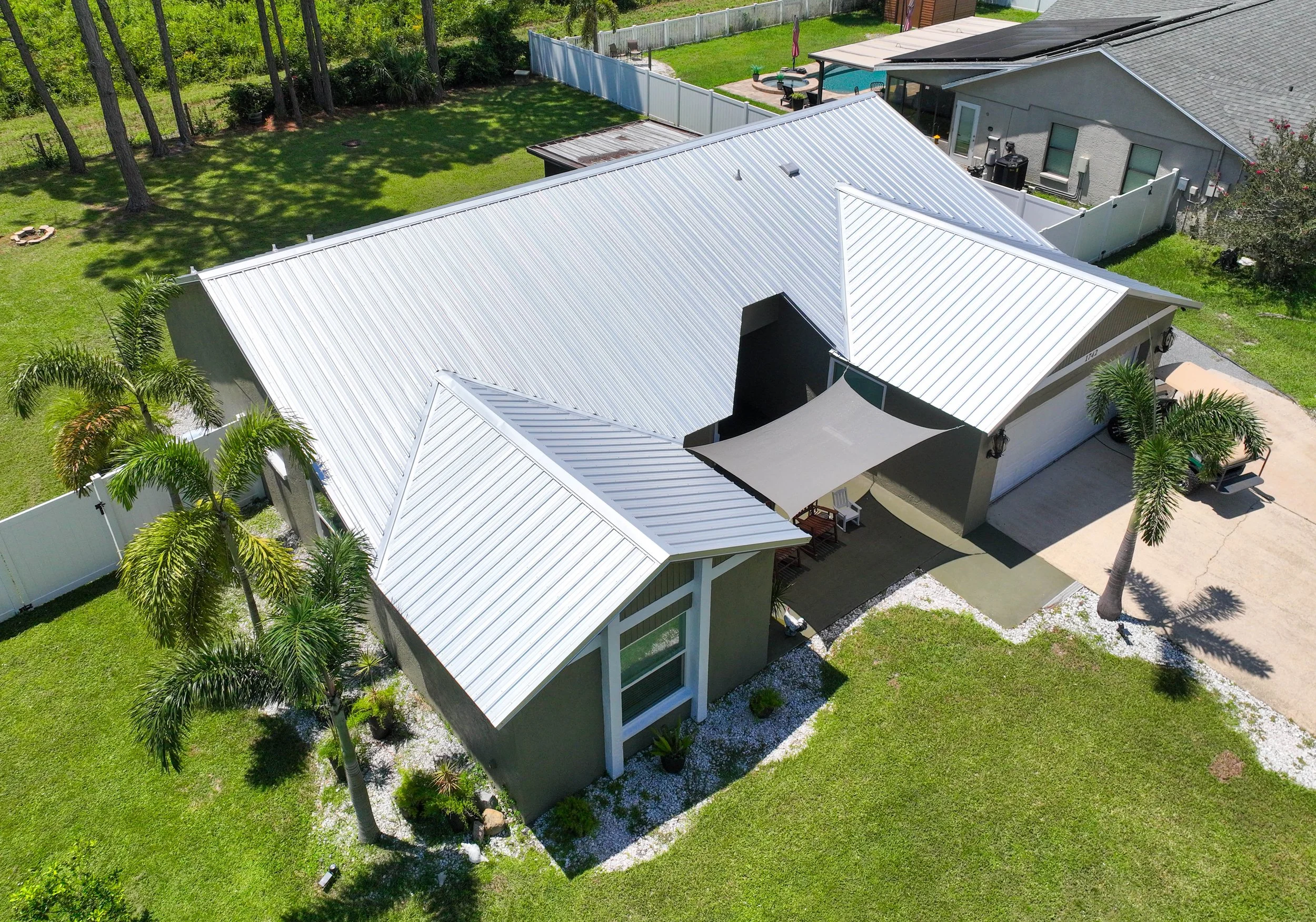 Aerial view of a house with a metal roof, surrounded by a green lawn, palm trees, and a white fence. There is a shaded patio area with outdoor furniture.