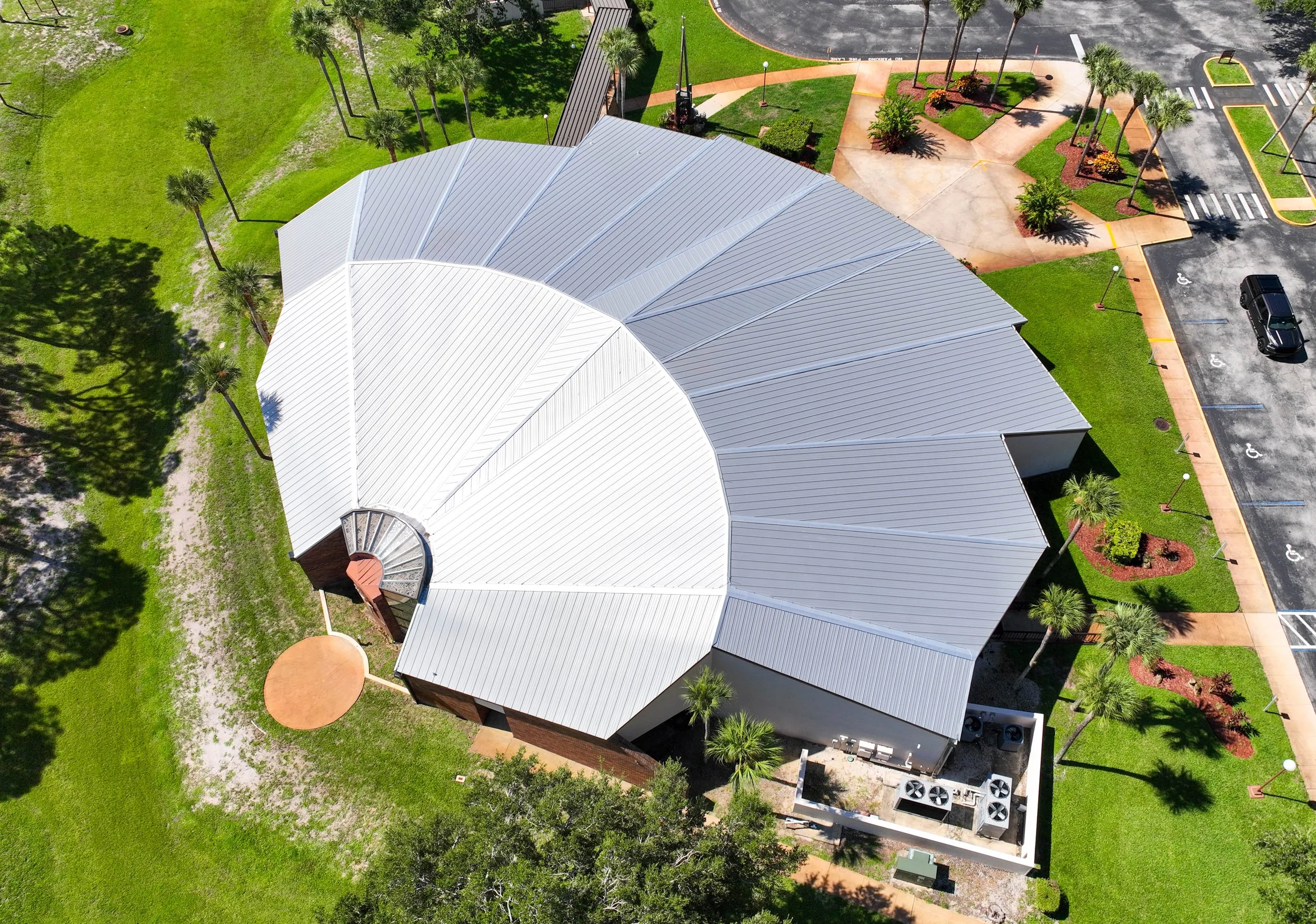 An aerial view of a building with a light-colored metal roof, surrounded by landscaped areas with palm trees, grass, and sidewalks, and parking lot with marked spaces.