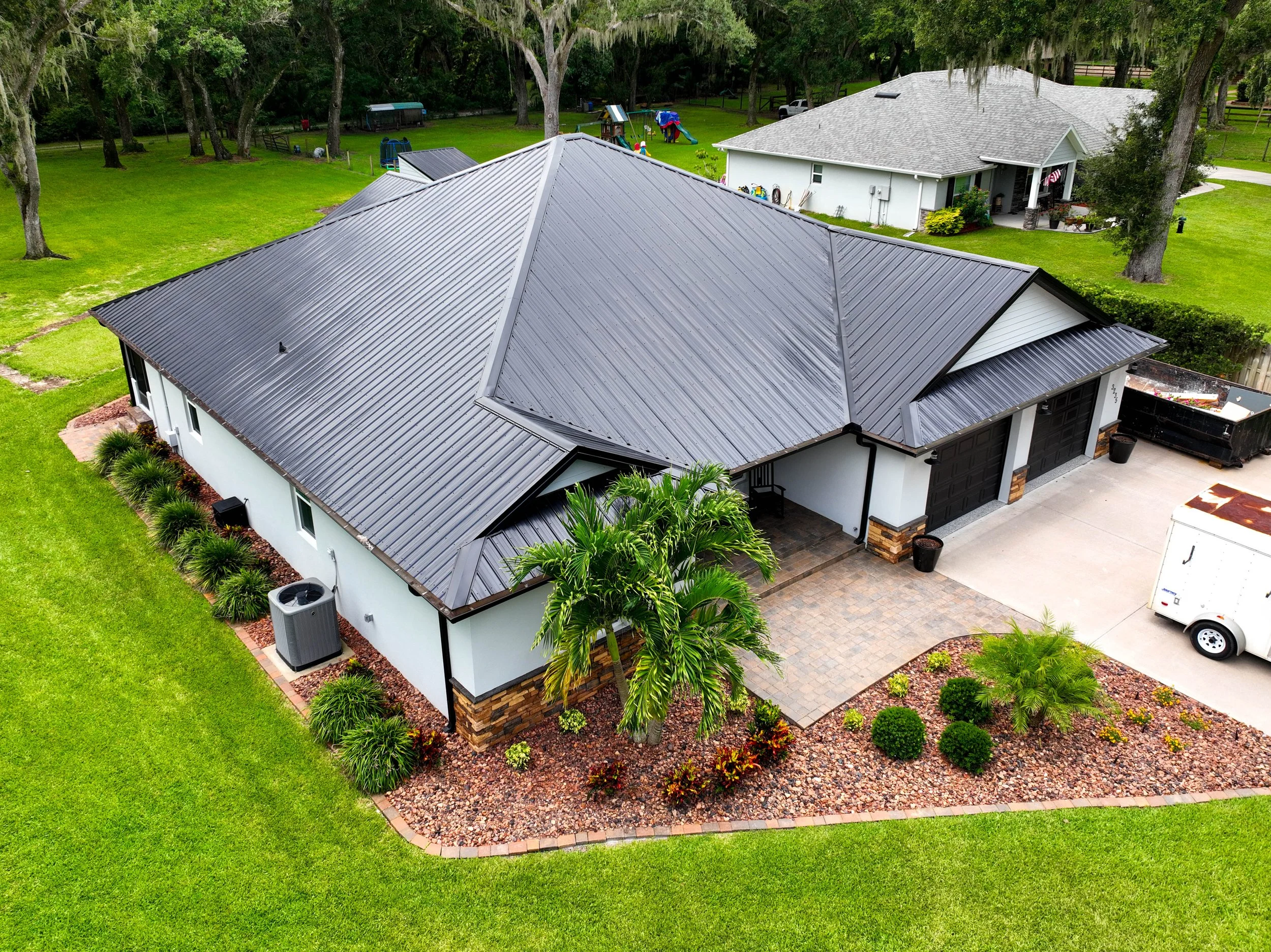 Aerial view of a house with a metal roof, surrounded by a well-manicured lawn, various plants, and trees, with a driveway and a white trailer nearby.