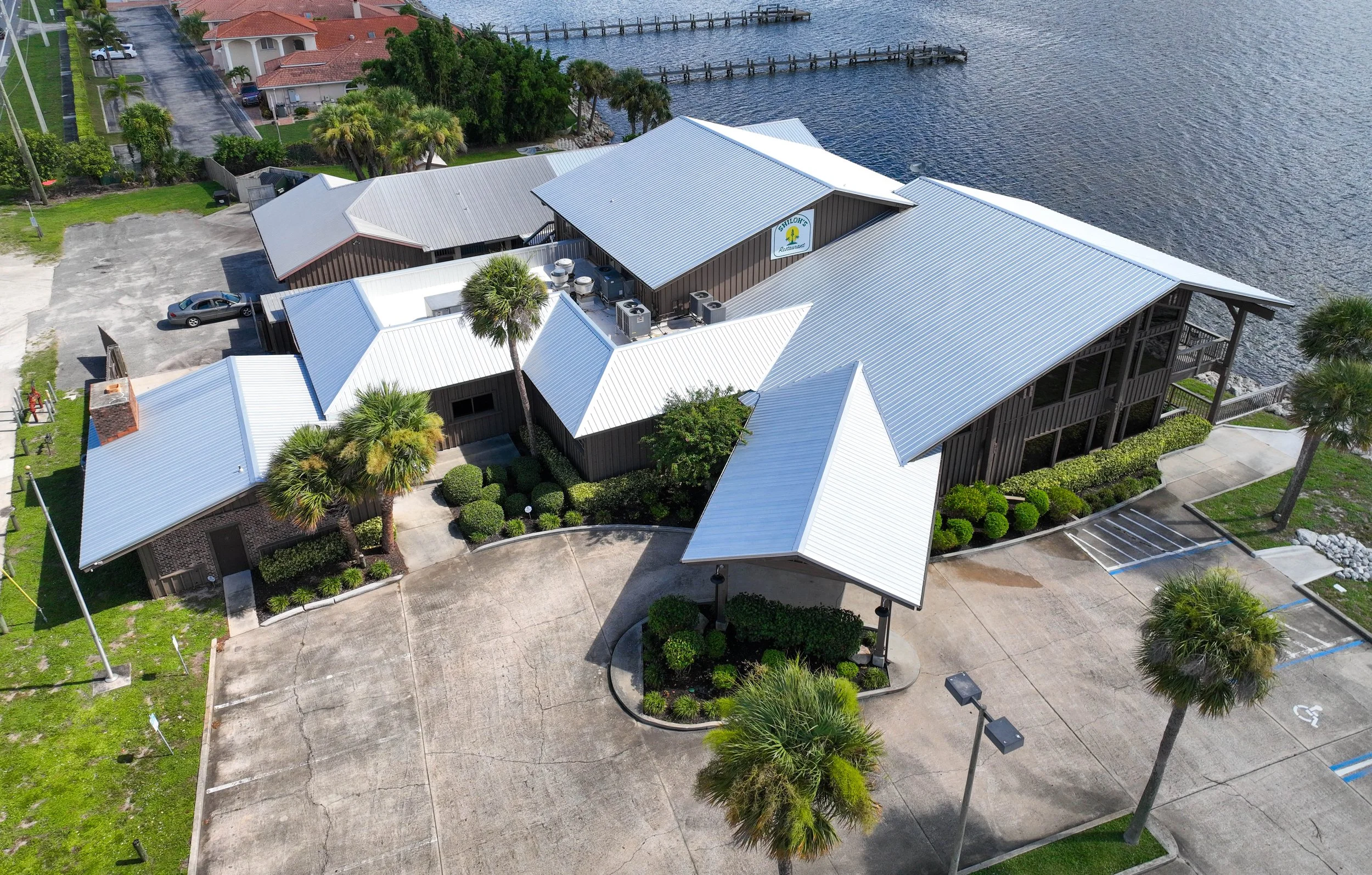 Aerial view of a waterfront building with metal roofs, surrounded by palm trees and a parking lot, near a body of water with a small pier.