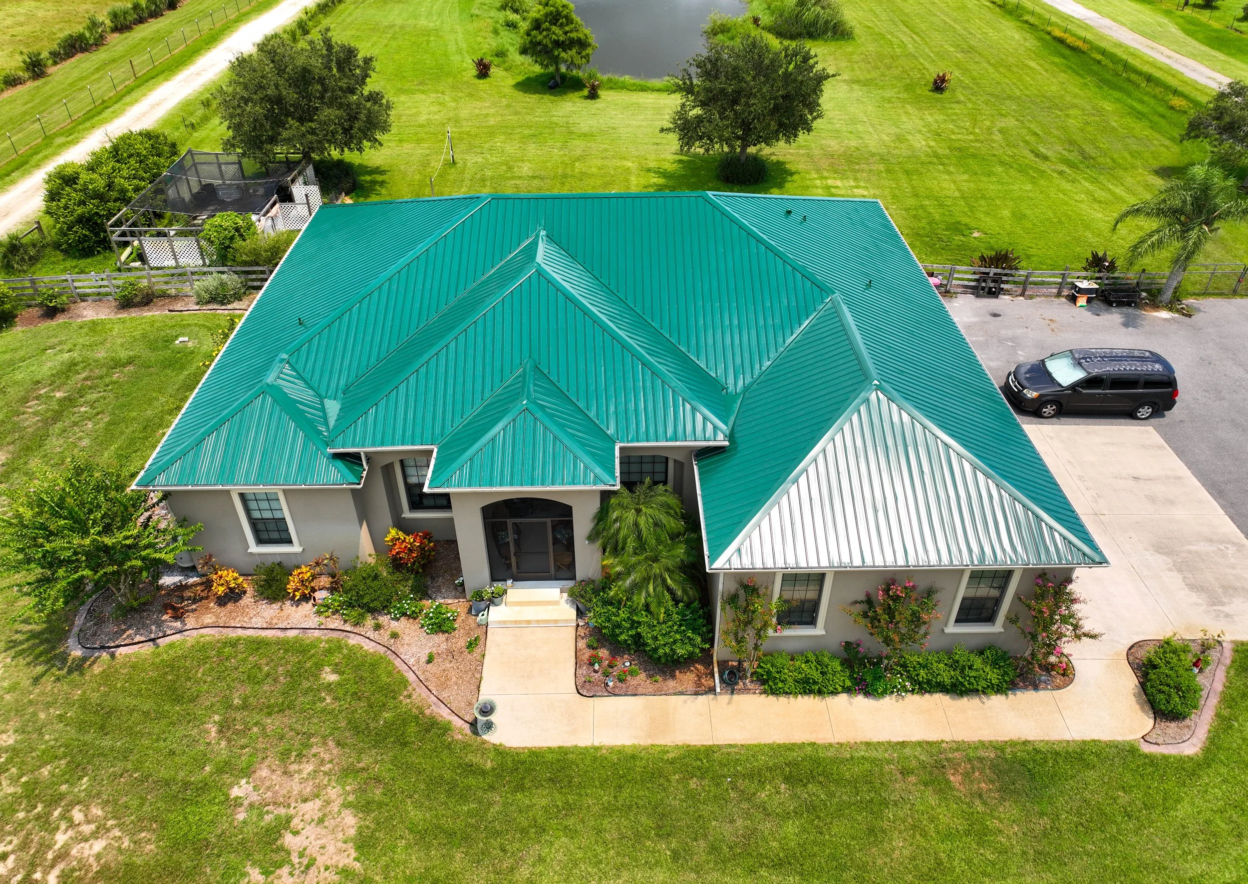 Aerial view of a single-story house with a teal metal roof, surrounded by a landscaped yard with various plants and trees, and a driveway with a black vehicle parked on it. There's a pond and green fields nearby.