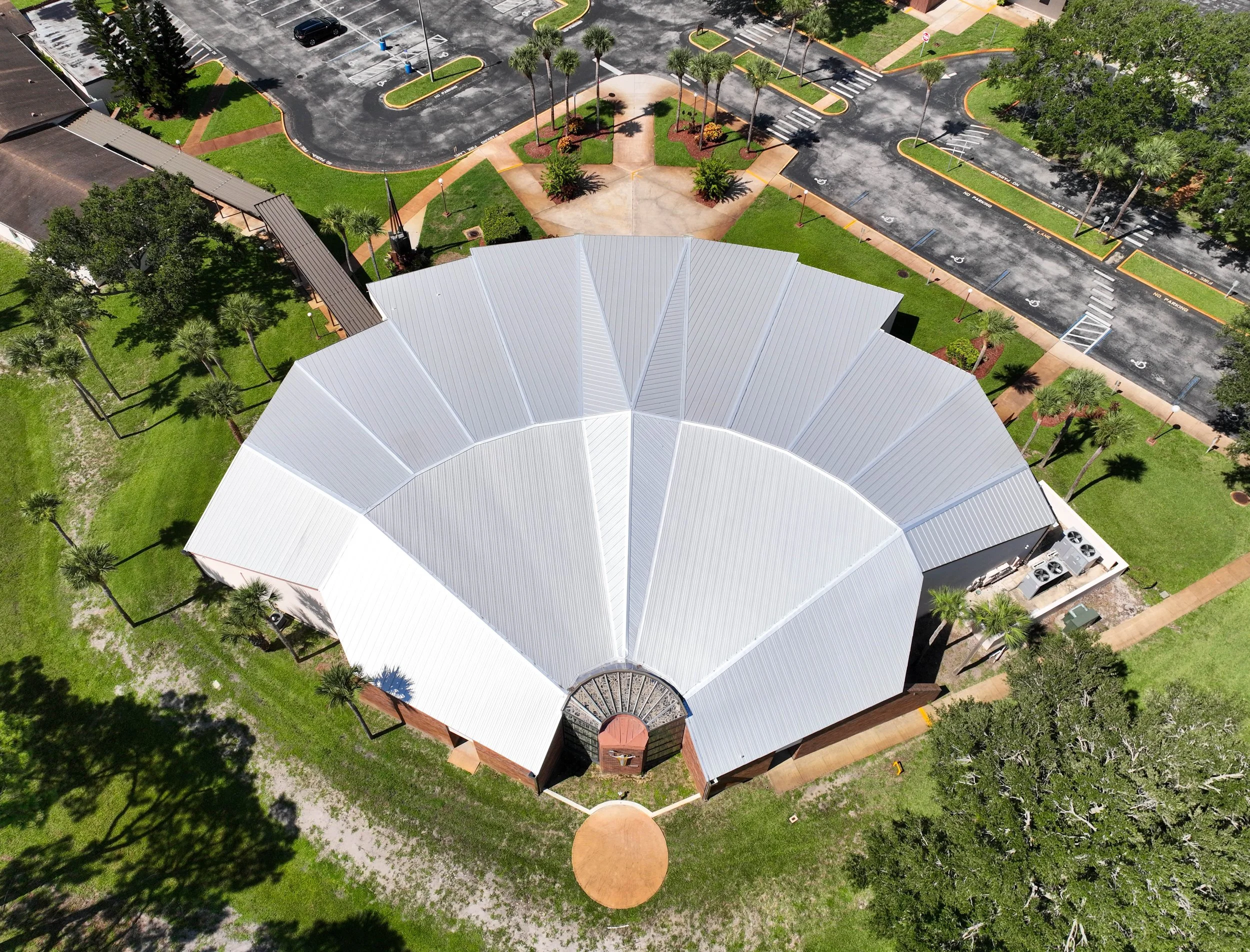 An aerial view of a circular building with a metal roof, surrounded by a green lawn, trees, and a parking lot with marked spots and Palm trees.