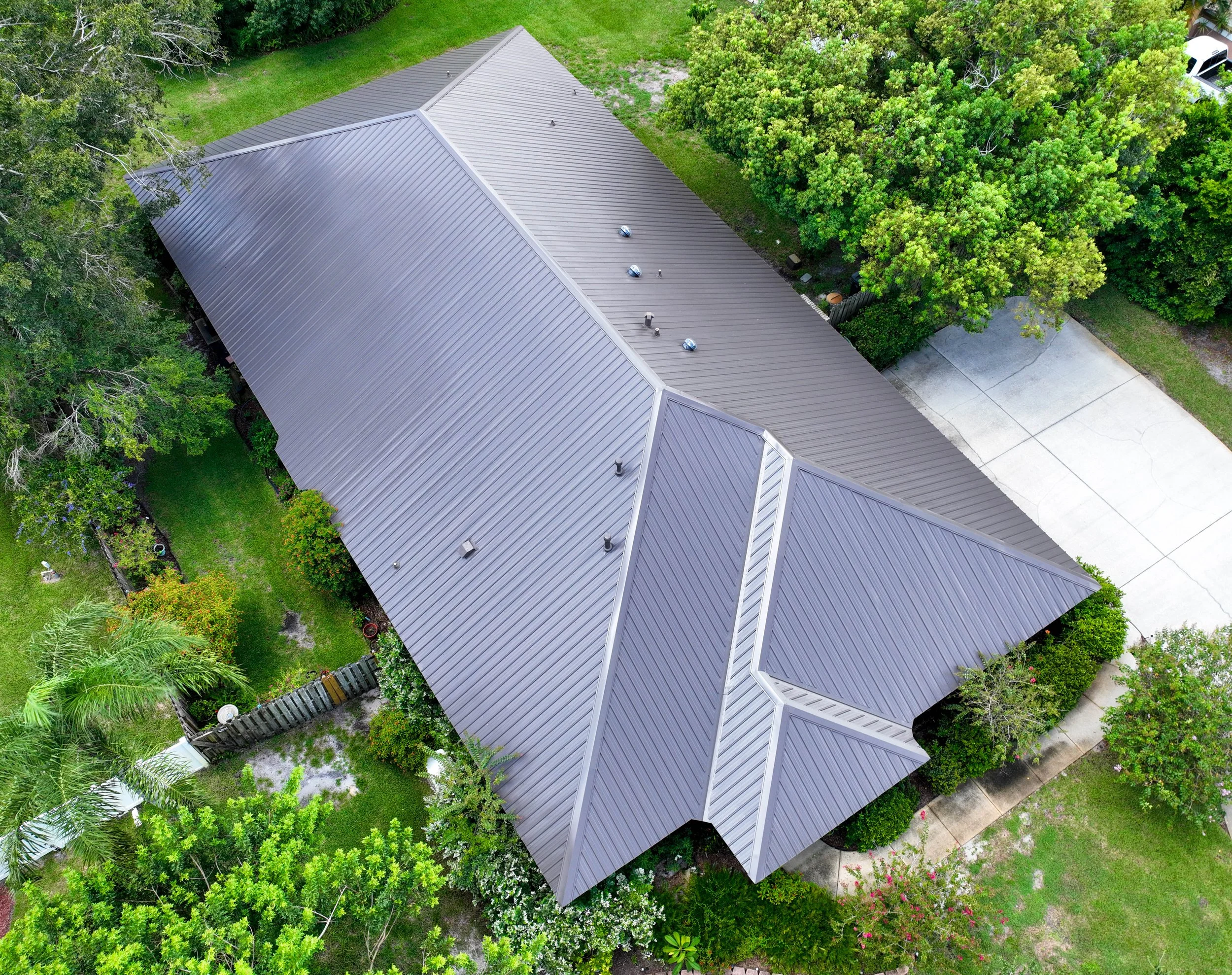 Aerial view of a house with a metal roof surrounded by green trees and a concrete driveway.