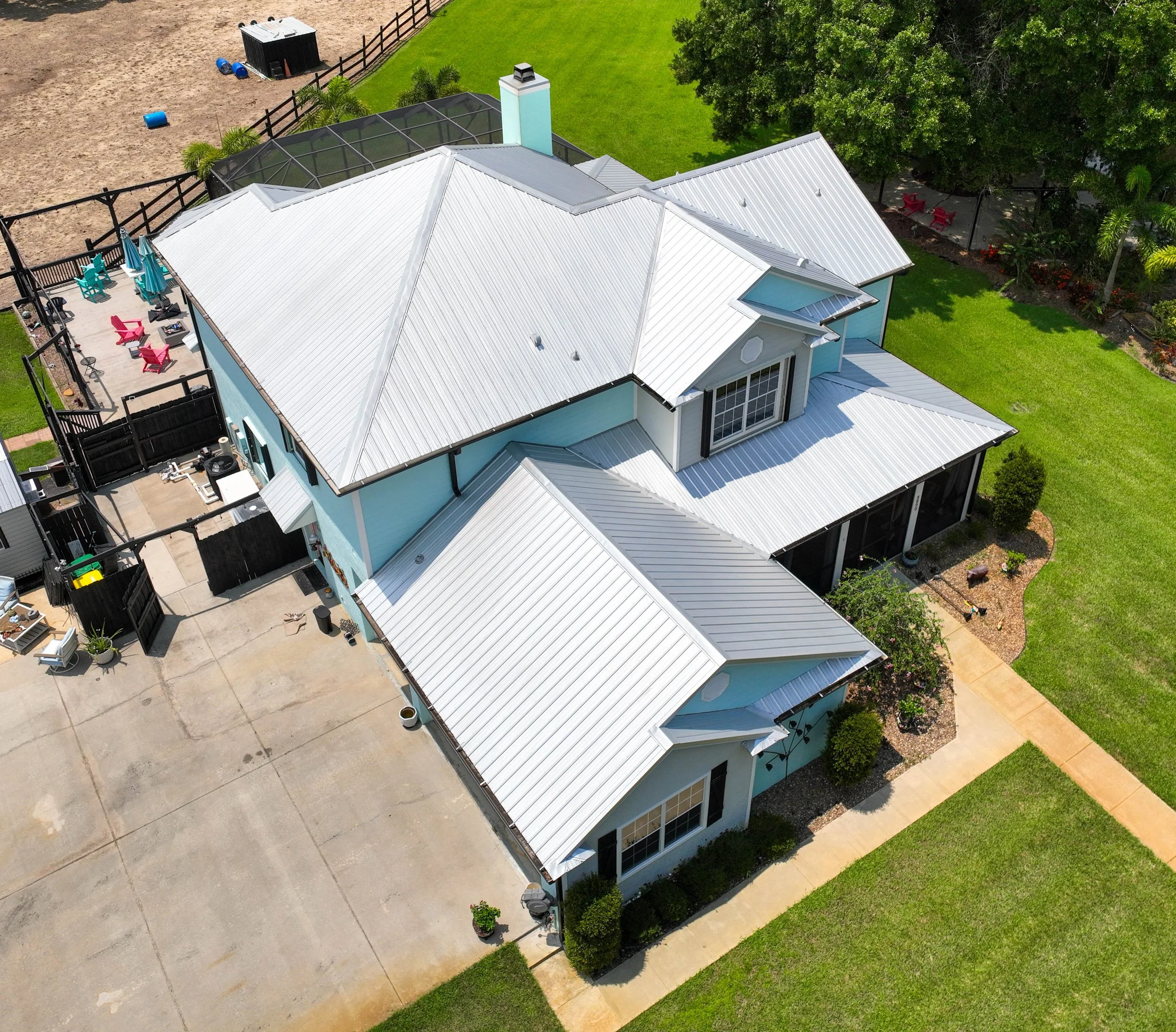 Aerial view of a house with a white metal roof, a concrete driveway, green lawn, and landscaping, including trees and bushes. There is a fenced backyard with patio furniture, and some outdoor storage structures.