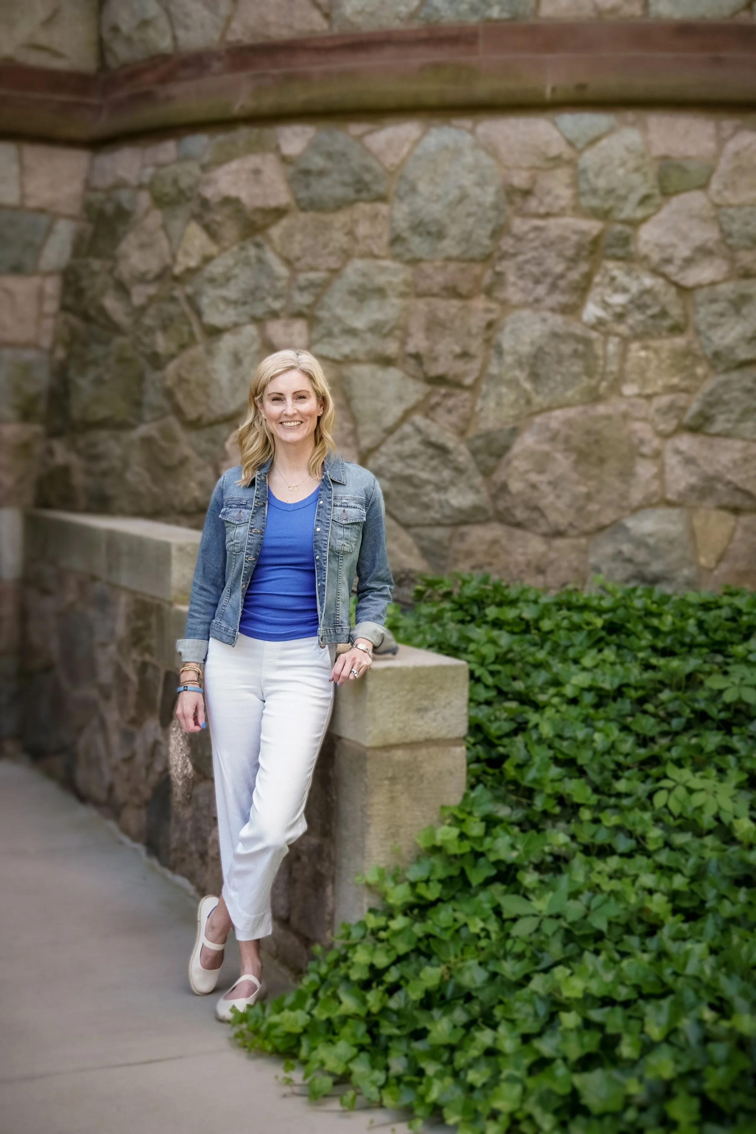 Smiling woman with blonde hair in a denim jacket and white pants standing by a stone wall and green bushes.