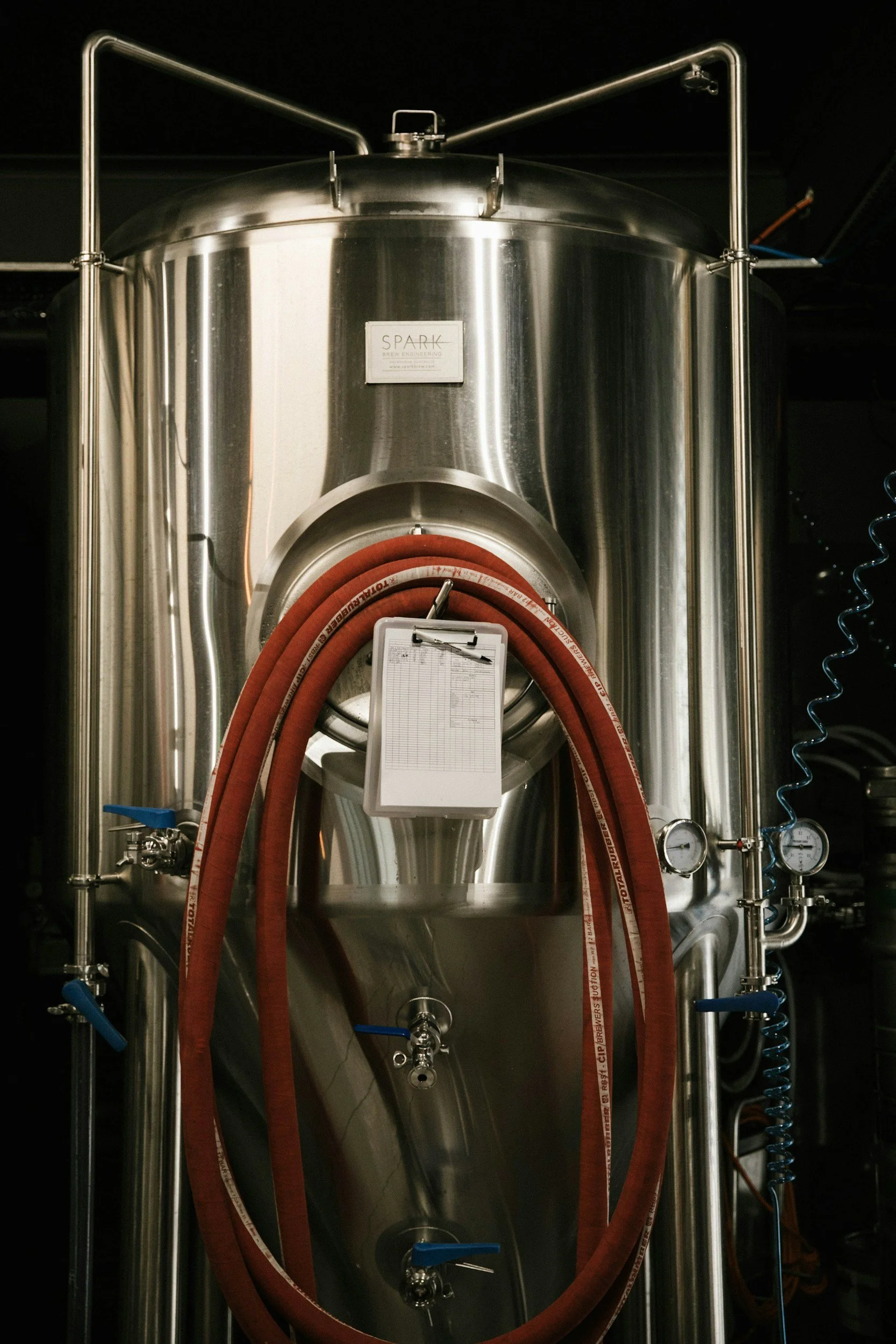 Large stainless steel brewing or processing tank with hoses, gauges, and a clipboard attached, in an industrial setting.