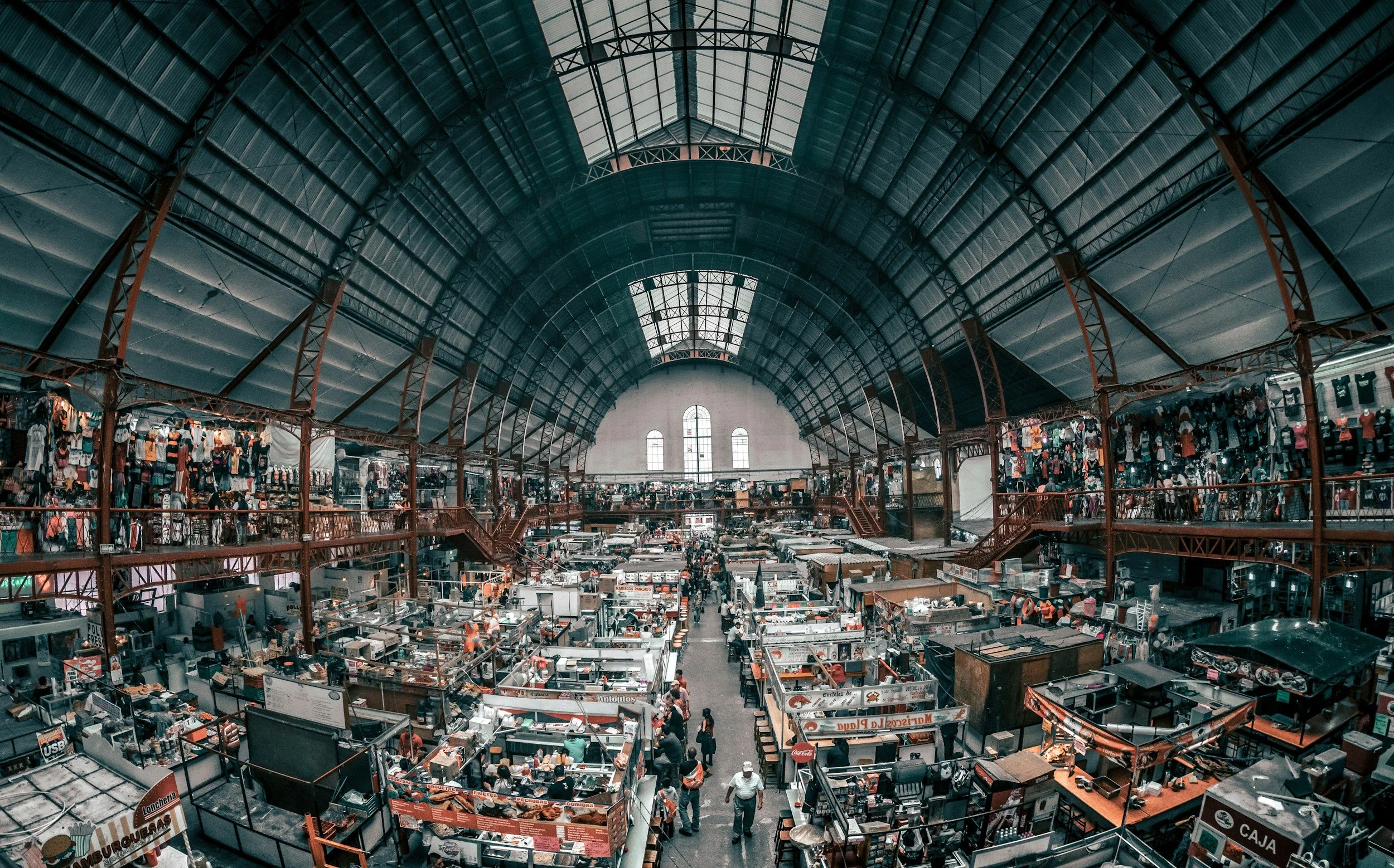 Interior of a large indoor market with high arched ceiling and multiple stalls selling various goods.