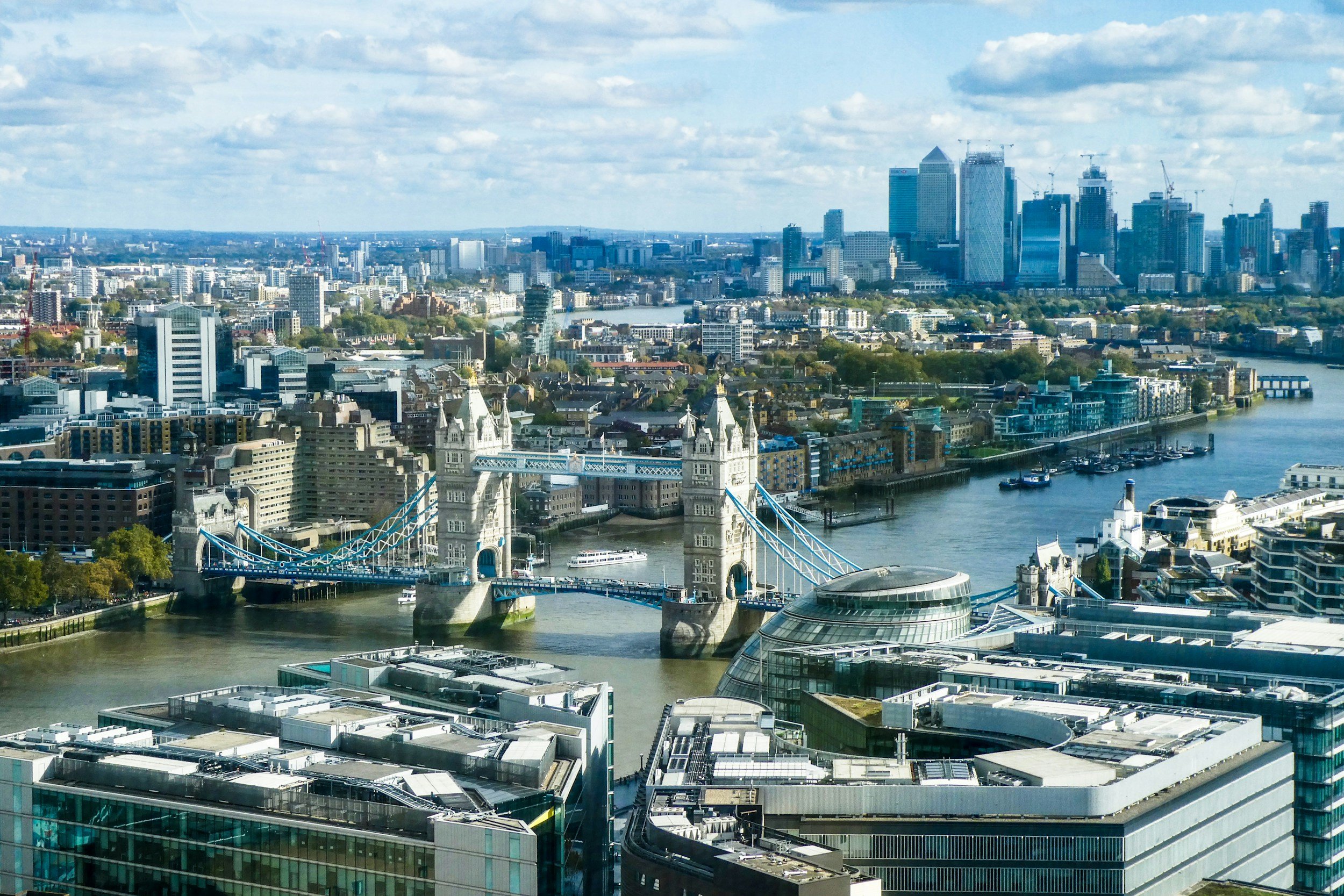 A panoramic view of London featuring Tower Bridge over the River Thames, with modern buildings and the city skyline in the background, under a partly cloudy sky.
