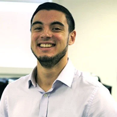 Smiling young man with dark hair and beard wearing a light-colored dress shirt in an office setting.