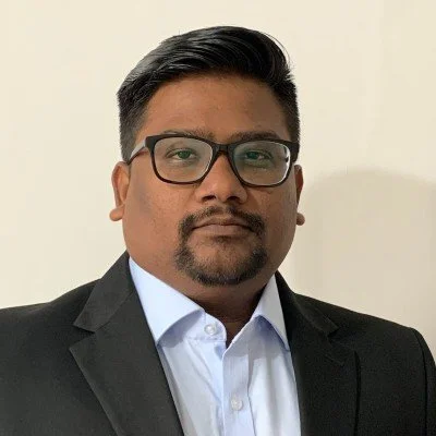 A man wearing black glasses and a black suit with a white shirt, standing against a plain light-colored background.