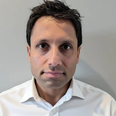 Close-up of a man with dark hair wearing a white dress shirt, facing the camera against a plain background.