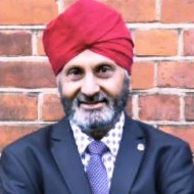 A man wearing a red turban, dark suit, and tie, smiling in front of a brick wall.