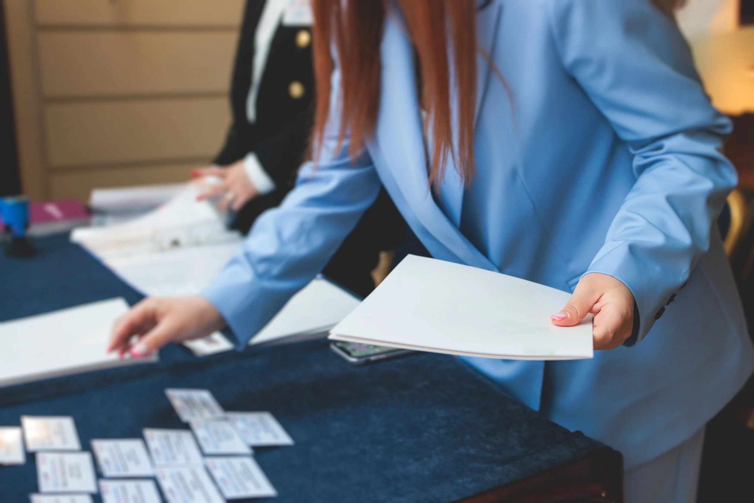 RSVP para eventos corporativos, pessoa pegando papeis ou documentos em uma mesa, com outros documentos espalhados na mesa, possivelmente em uma reunião ou conferência.