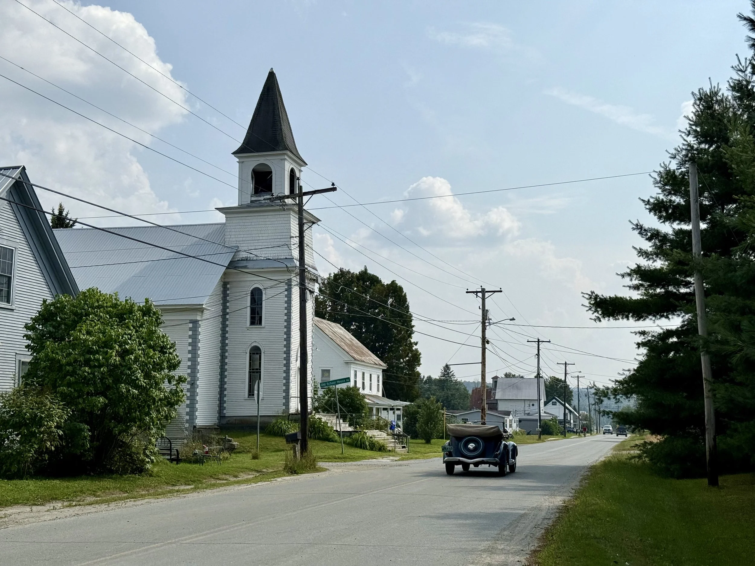A small town street scene with a white church in Greensboro Bend, VTfeaturing a tall steeple, houses, a tree-lined street, utility poles and wires, and a golf cart on the road on a sunny day with some clouds in the sky.