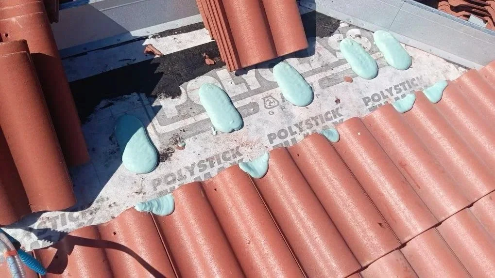 Roof with orange clay tiles, green foam sealant applied in several spots, metal sheathing with printed text, and shadows from nearby objects.