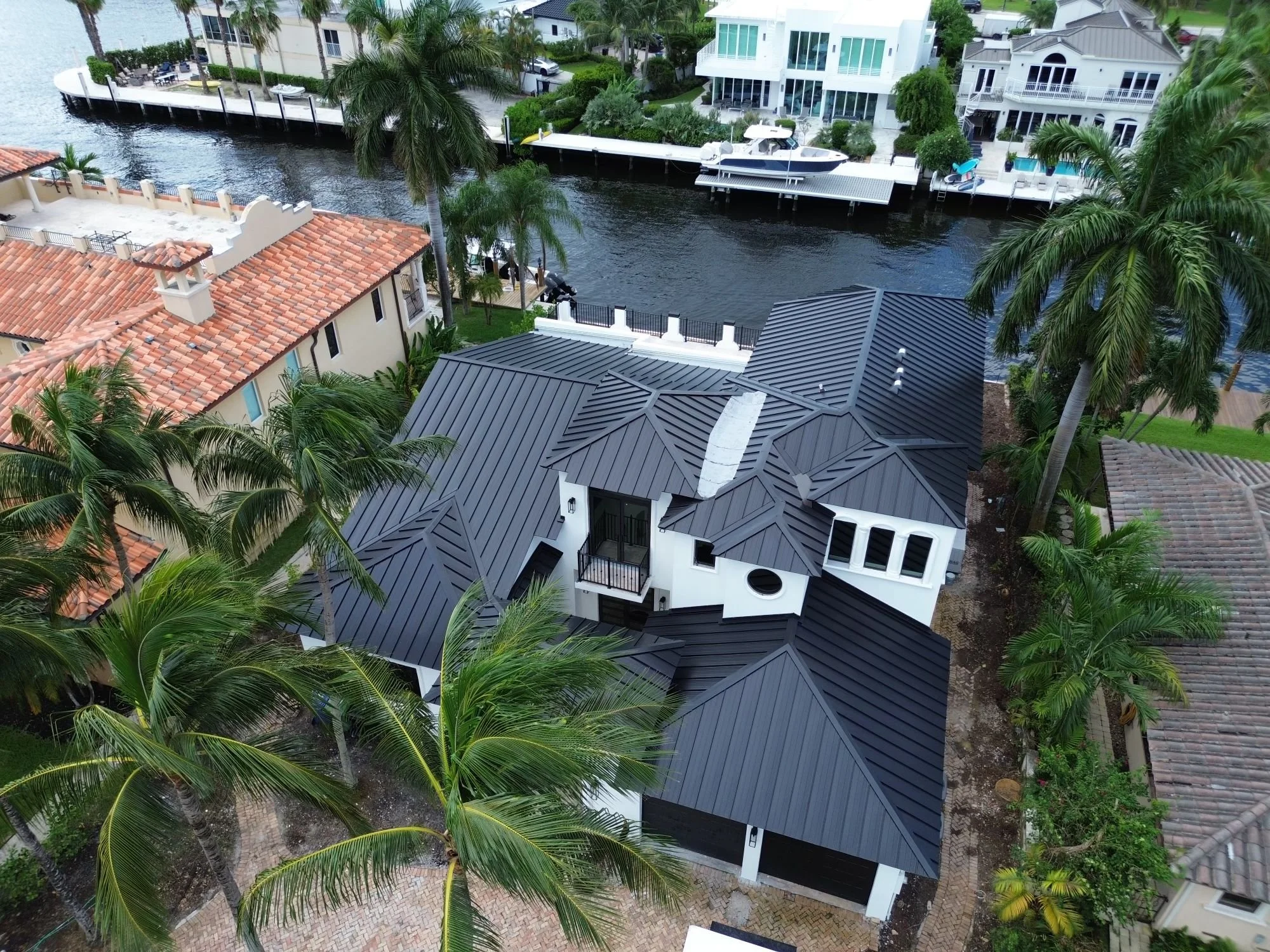 An aerial view of a modern house with a black metal roof, surrounded by palm trees, neighboring houses, and a canal with boats in a waterfront neighborhood.