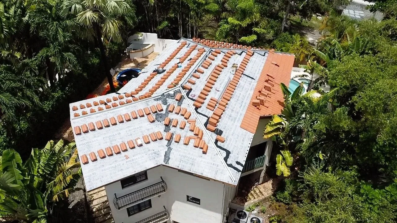 Aerial view of a house with a partially completed roof renovation, showing new clay tiles being installed on sections of the roof with some areas still covered with roofing underlayment.