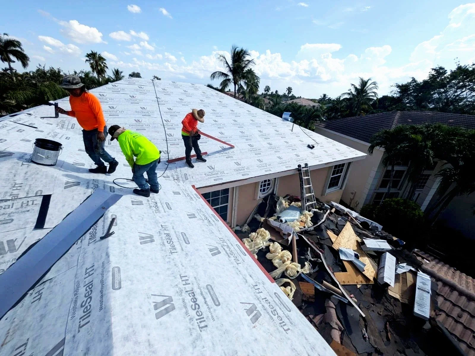 Construction workers installing a new roof on a house, with tools and materials visible, surrounded by palm trees in a sunny environment.