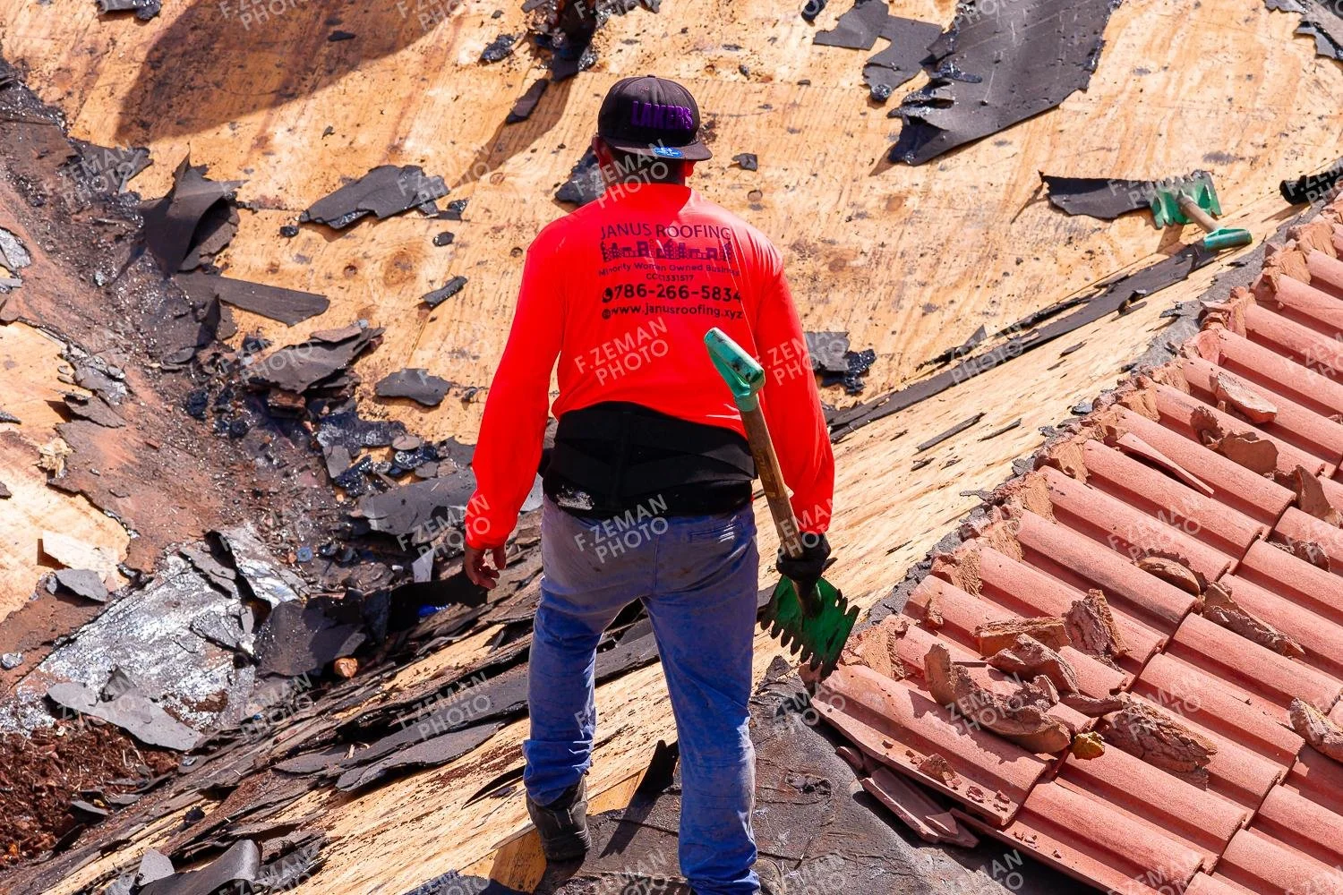 A construction worker standing on a roof with damaged tiles and debris, holding a green tool.