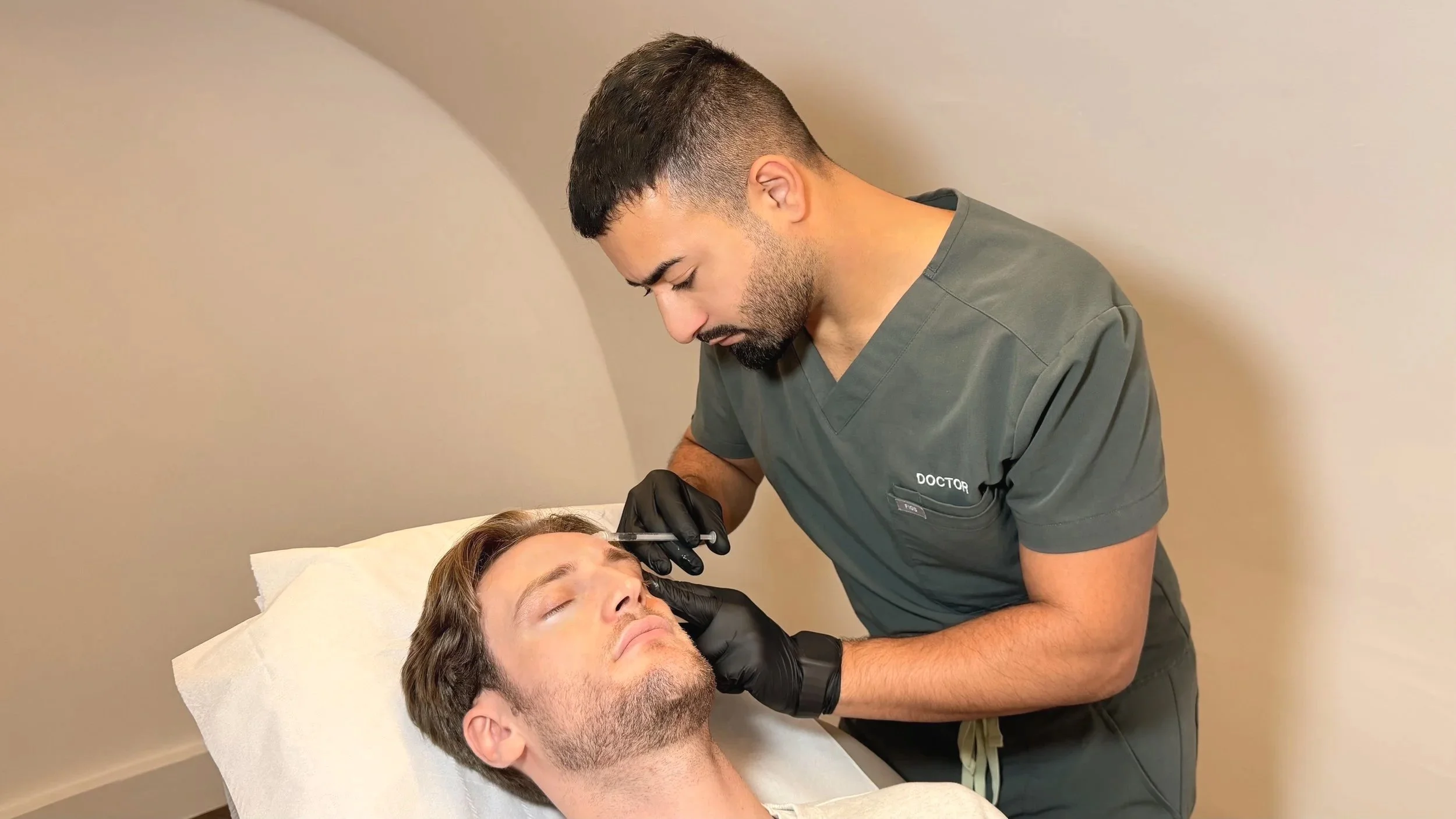 A male doctor in scrubs and black gloves performs a cosmetic procedure on a male patient lying in a medical bed with closed eyes.