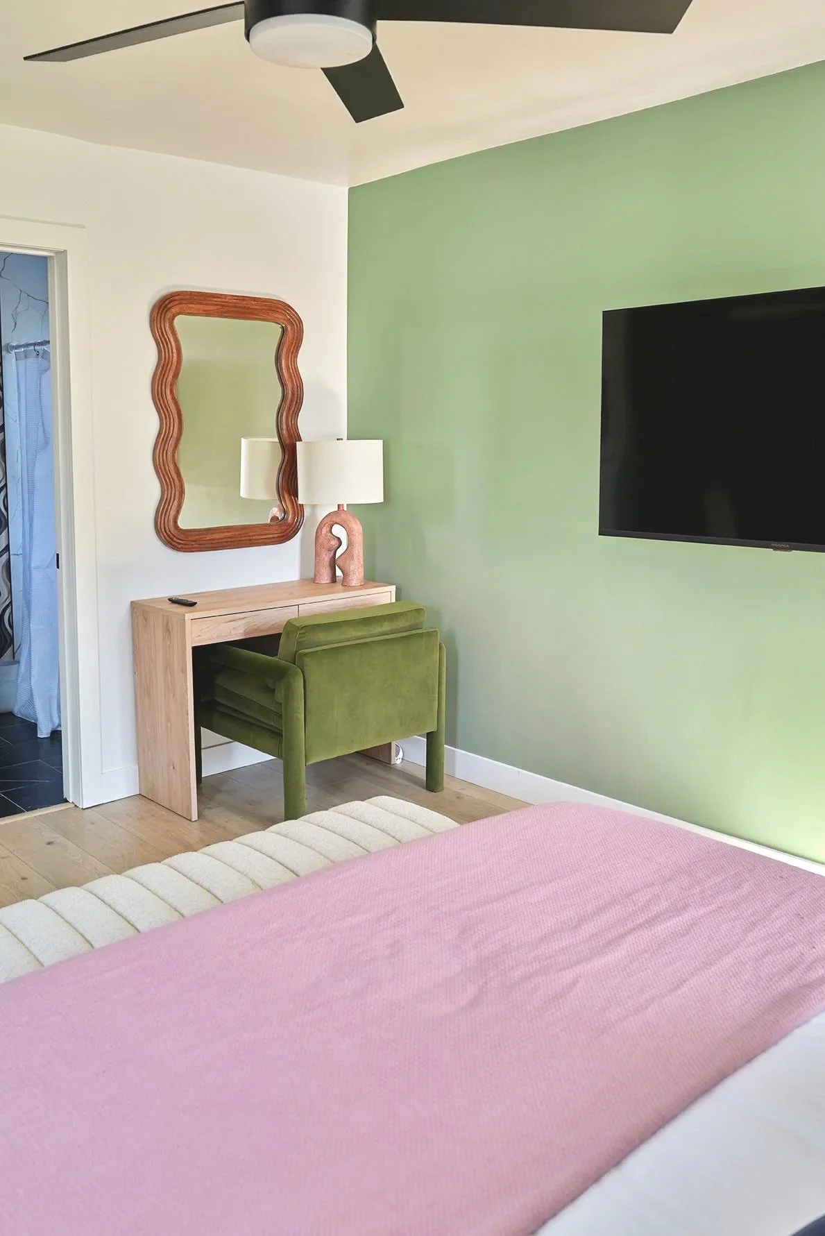 A bedroom corner with a green accent wall, a wall-mounted flat-screen TV, a small wooden desk with a green chair, a wavy-edge mirror, a table lamp with a textured base, and part of a bed with a pink blanket.
