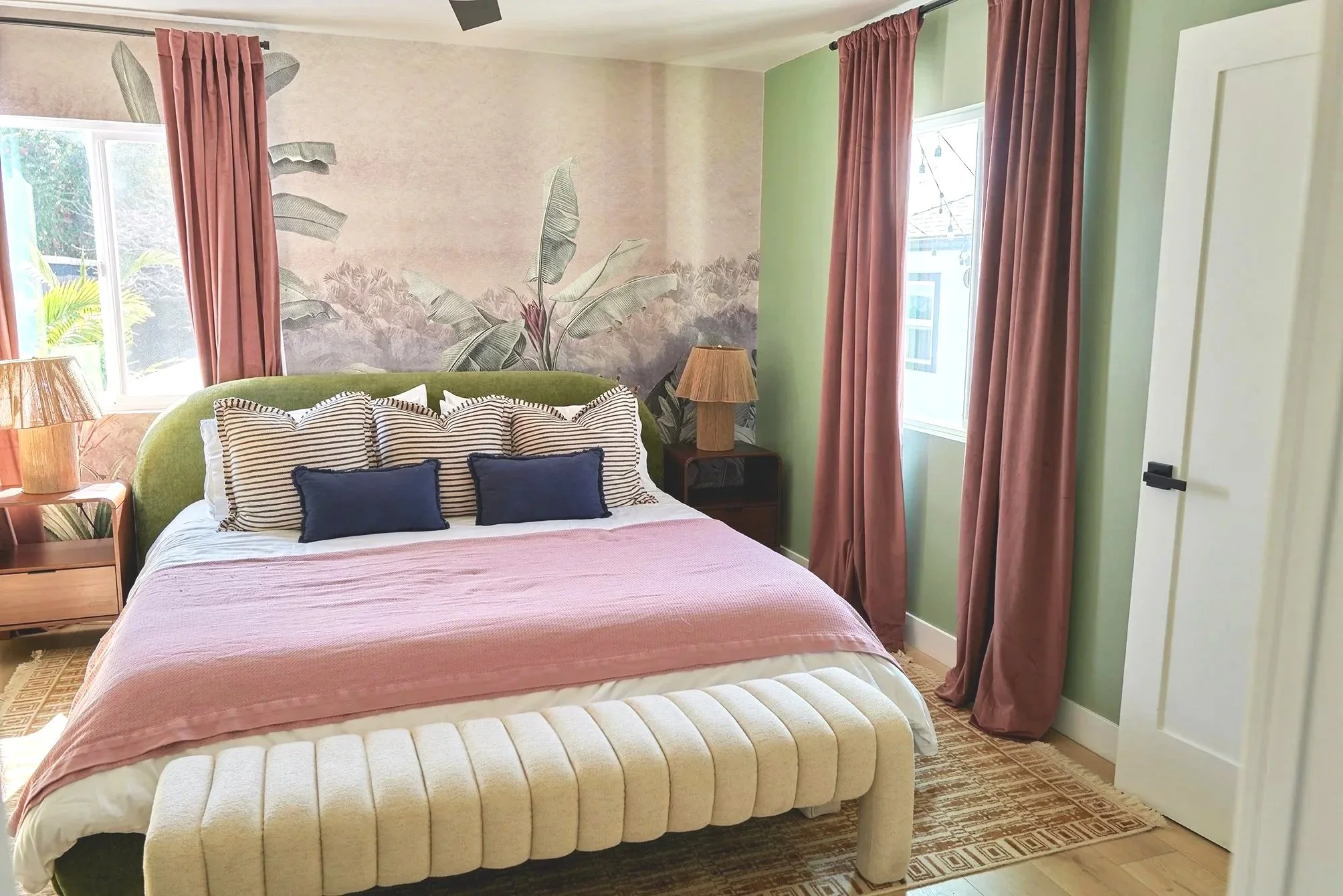 A bedroom with a bed that has a green upholstered headboard, striped pillows, navy pillows, and a pink blanket. There are pink curtains on the windows, a nightstand with a lamp, and a wall mural of tropical plants behind the bed.