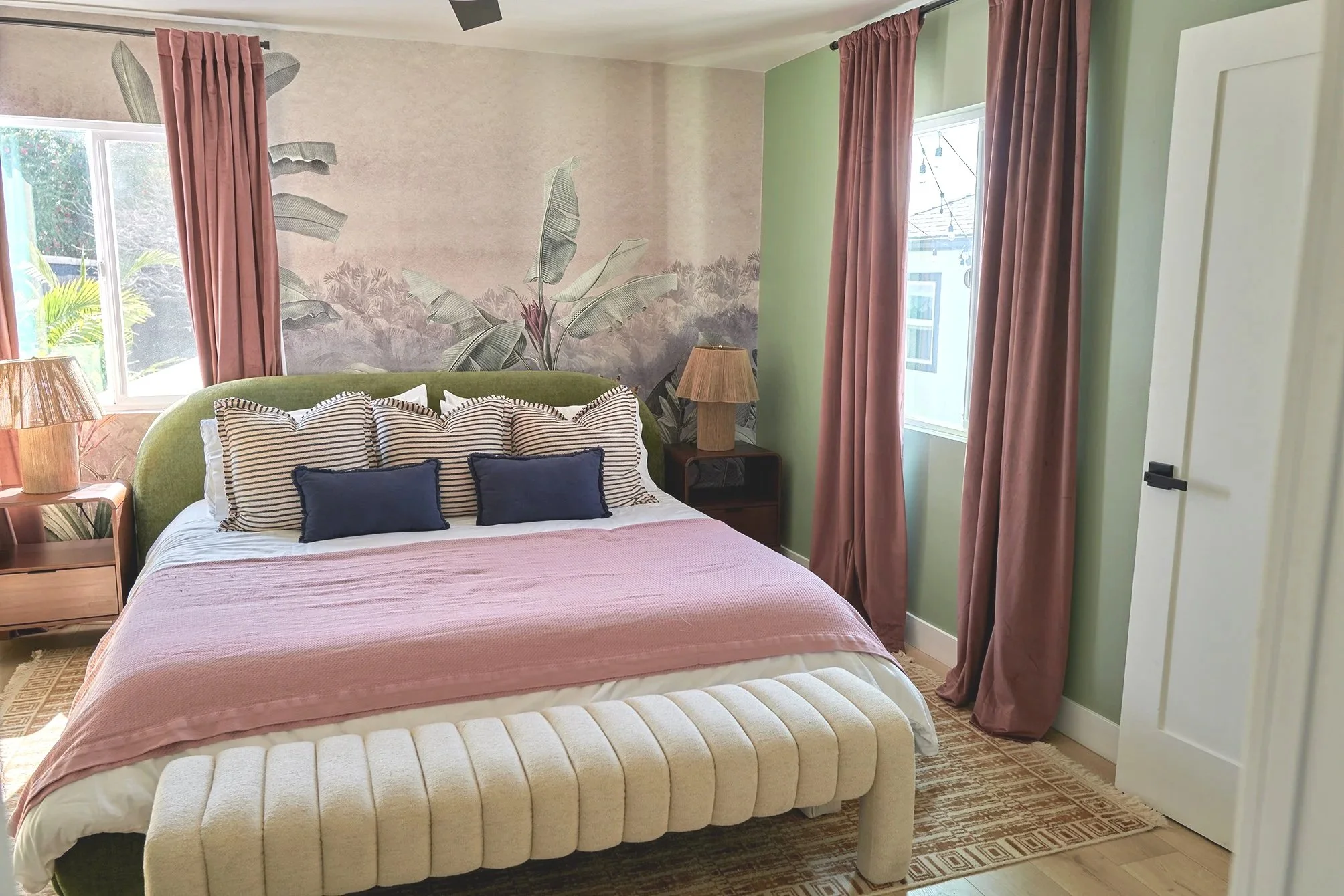 A bedroom with a green and pastel color scheme, featuring a bed with striped and navy pillows, pink bedding, and a velvet headboard. Pink curtains hang on two windows, and there is a nightstand with a tan lampshade on one side. A mural with tropical 