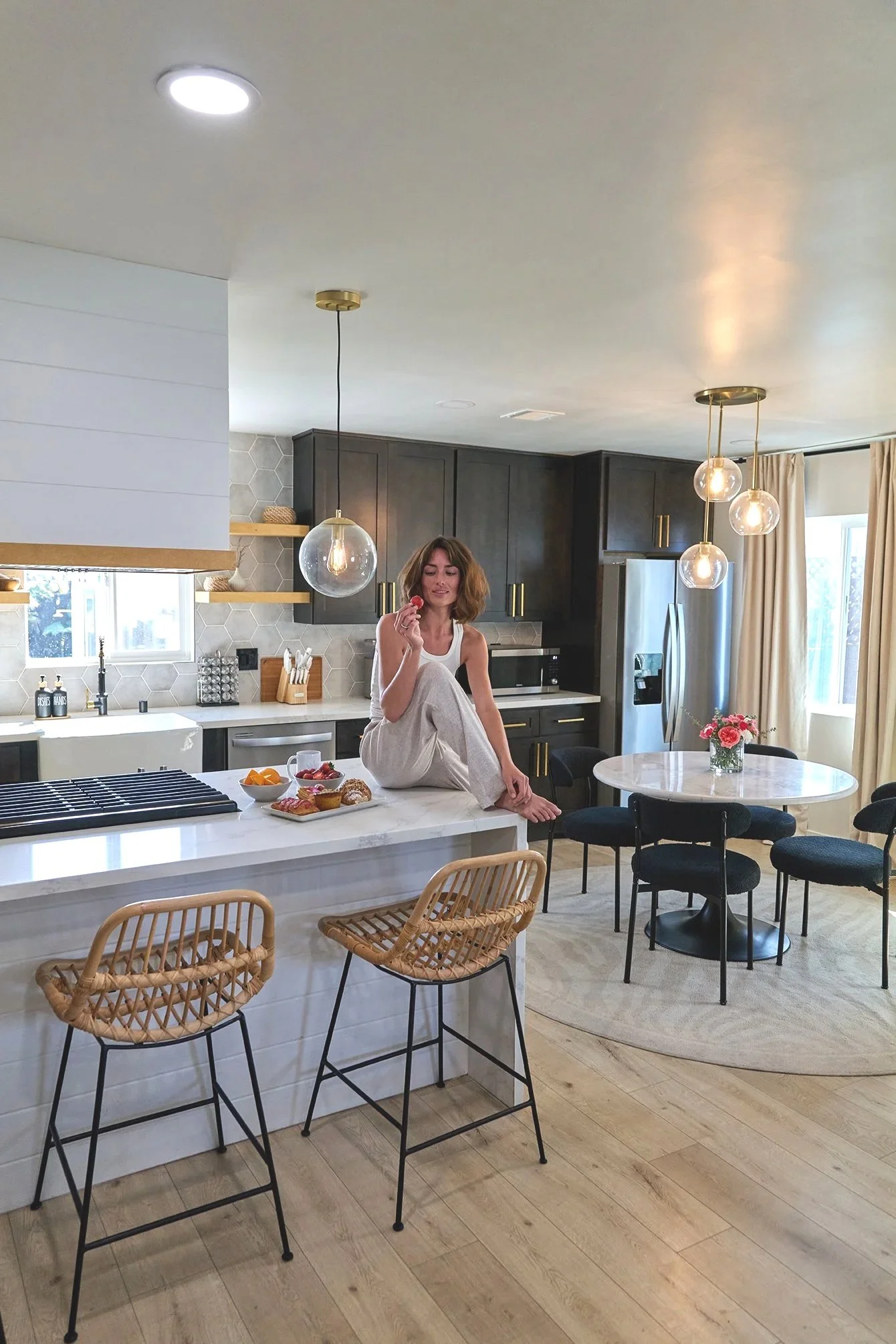 A woman sitting on a kitchen island eating a strawberry with a bowl of fruit and baked goods on the counter. The kitchen features dark cabinets, a round dining table with black chairs, and modern hanging lights, with natural sunlight coming through w