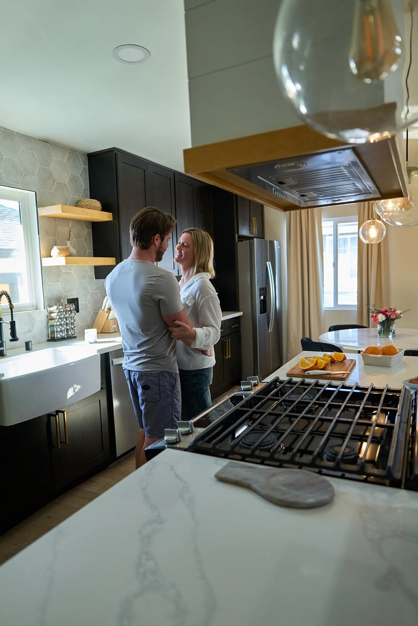 A couple standing close together and smiling in a modern kitchen with black cabinets, white countertops, and a window, with a round table and a vase with flowers in the background.