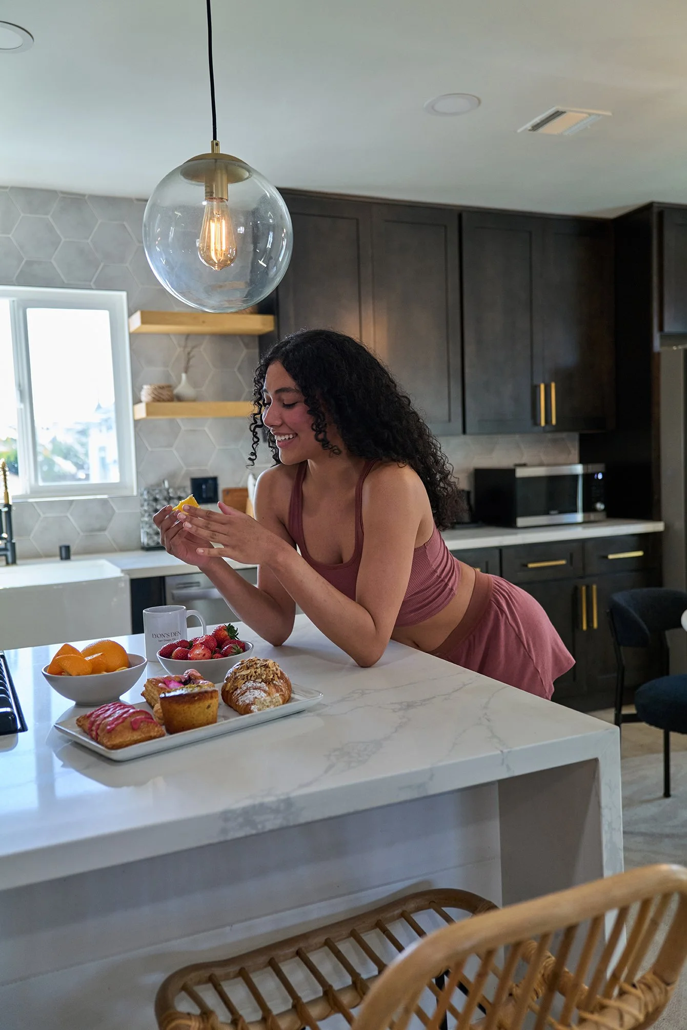 A young woman in a pink workout outfit leaning on a white marble kitchen island, smiling while holding a lemon, surrounded by bowls of strawberries, oranges, and baked goods in a modern kitchen.