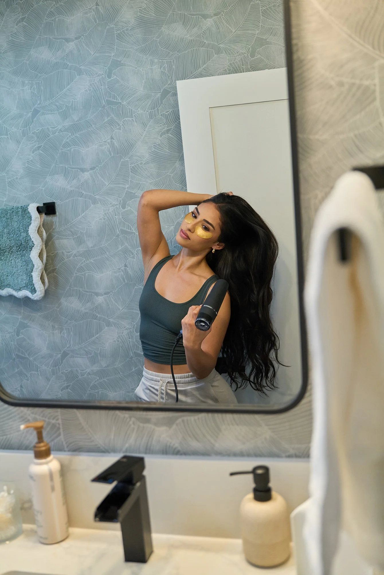 A woman taking a mirror selfie in a bathroom, holding a hairdryer in one hand and with gold under-eye masks, with long dark wavy hair, wearing a dark green tank top and light-colored shorts.