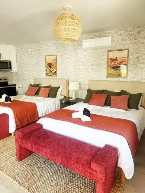 Two beds in a hotel room with white bedding, red bed runners, and pillows in black and red. A red bench at the foot of the bed, a nightstand with a white lamp, and framed artwork on the textured wall. A hanging wicker light fixture and an air conditi