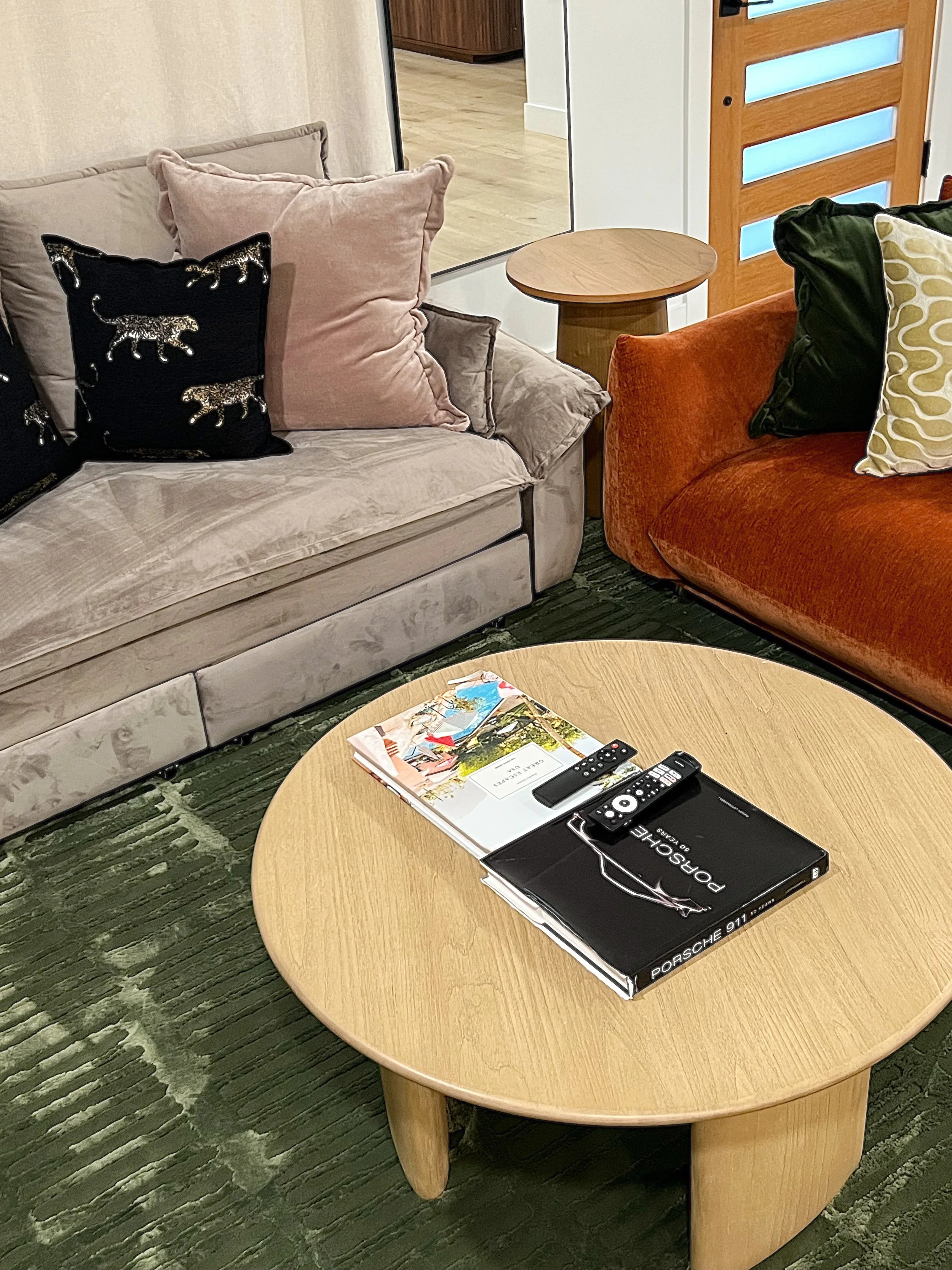 A living room with two sofas, one beige and one orange, with decorative pillows. A round wooden coffee table holds magazines, remotes, and a black book. A small wooden side table is near the beige sofa. The room has a green patterned carpet and a woo