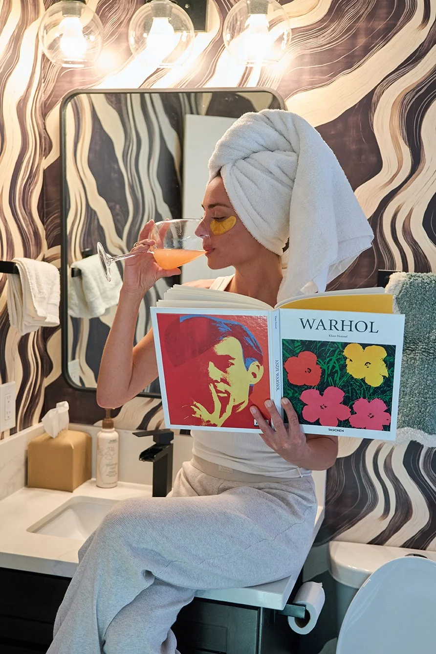 A woman with a towel wrapped around her head, wearing a white tank top, sitting on a bathroom counter. She is reading a large book about Warhol, holding a cocktail in one hand, and has yellow facial masks under her eyes. The bathroom has black and wh