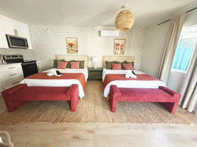 Two beds with red benches at the foot of each, white bedding, and pink and green pillows in a bright room with artwork on the textured wall, a hanging light, and a window with curtains.