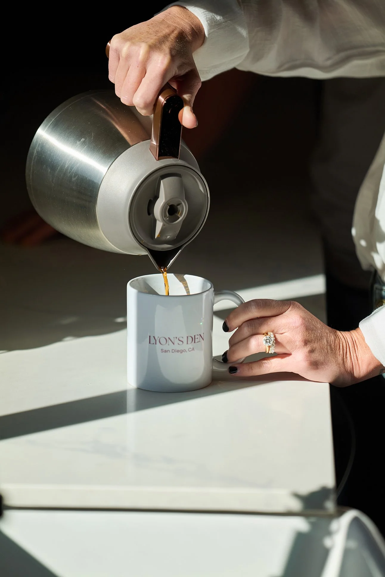 Person pouring coffee from a thermal carafe into a white mug labeled 'LYON'S DEN San Diego, CA' with sunlight casting shadows on the surface.