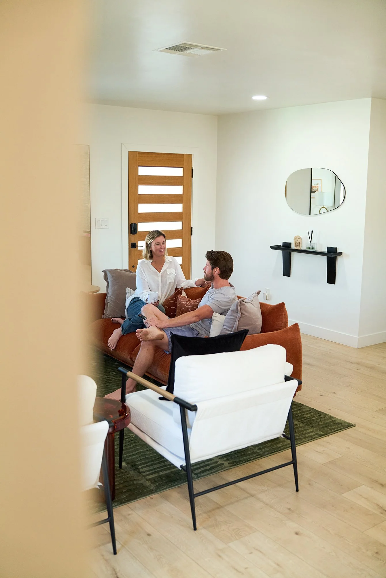 A couple sitting on a brown sofa in a modern living room, engaging in conversation. The room has a wooden door, a mirror on the wall, and minimalist decor. Part of a white chair is visible in the foreground.