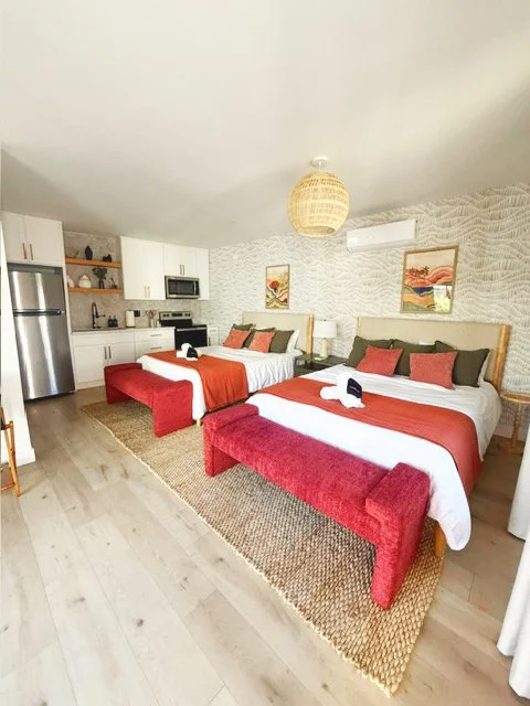 Two beds with red and black bedding in a modern studio apartment with kitchen, artwork, and decor.