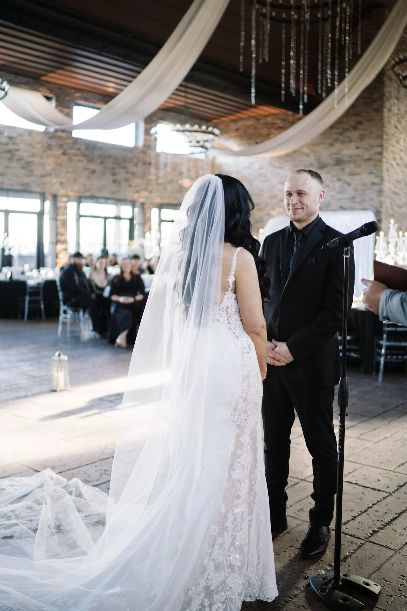 A bride and groom exchanging vows during their wedding ceremony inside a rustic venue with exposed brick walls, draped fabric decorations, and large windows.