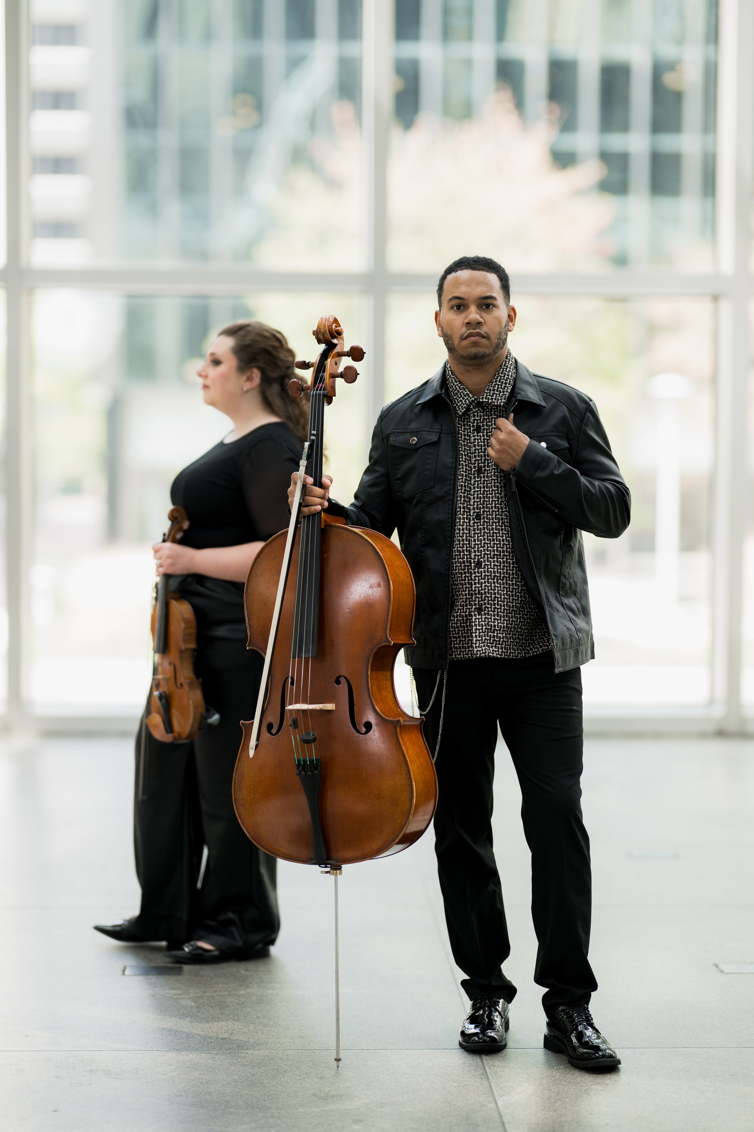Nicole Lennartz holding her violin and Ahmer'e Blackman holding his cello, standing in front of large glass window in a modern building.