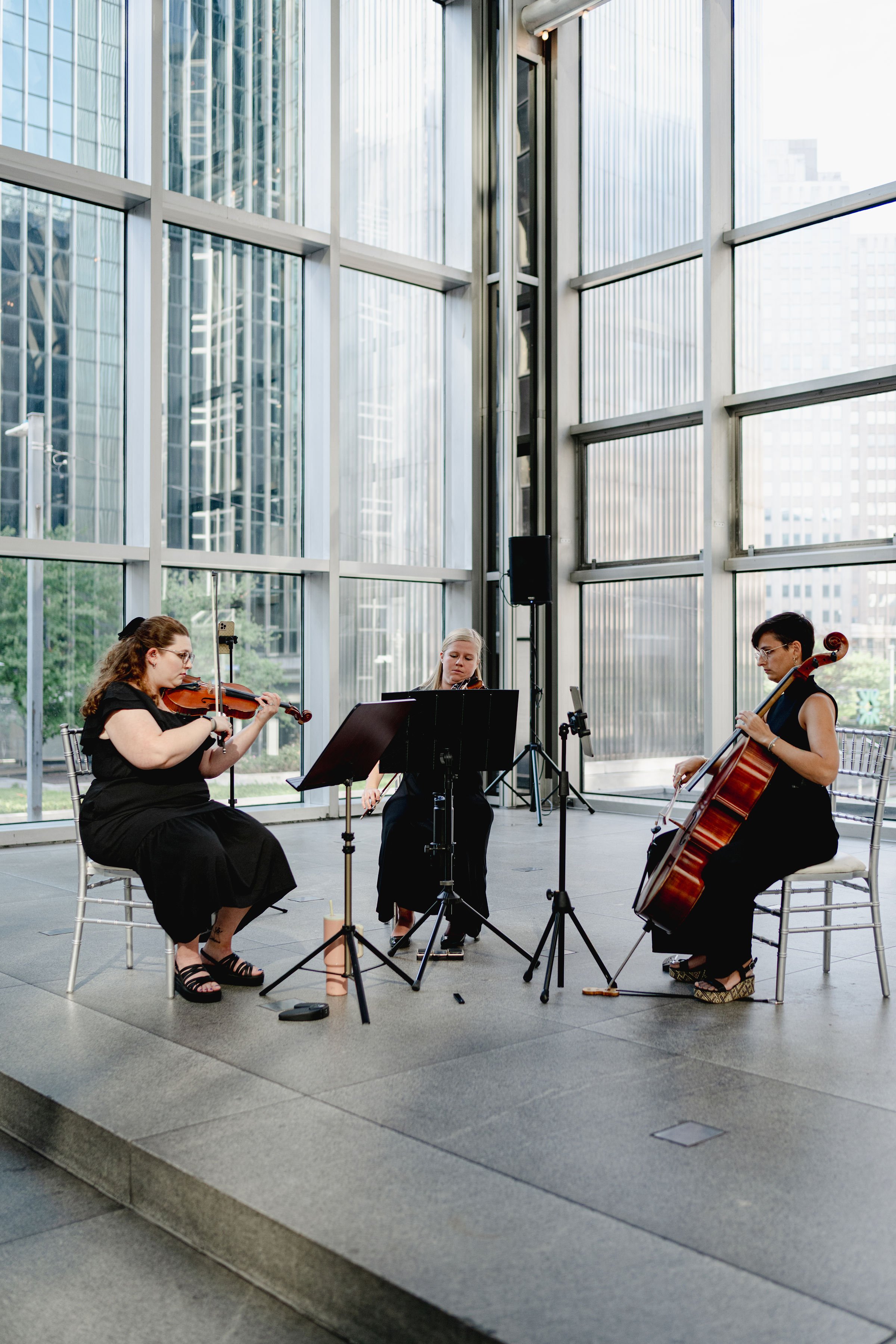 Radiant Strings trio playing violin, viola, and cello inside a modern glass building with cityscape view.