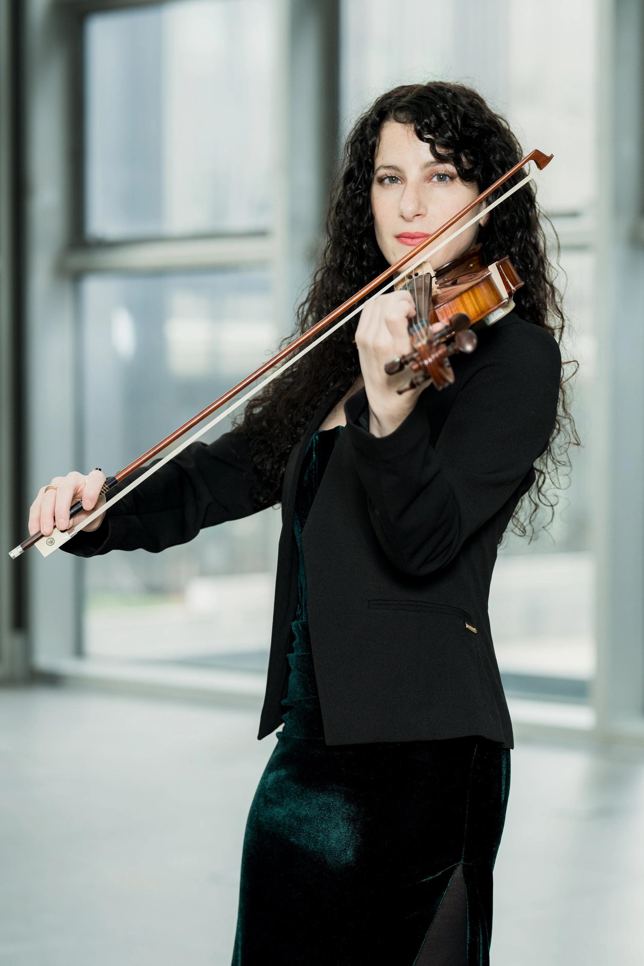 Cara Garofalo playing a violin indoors with large windows behind her.