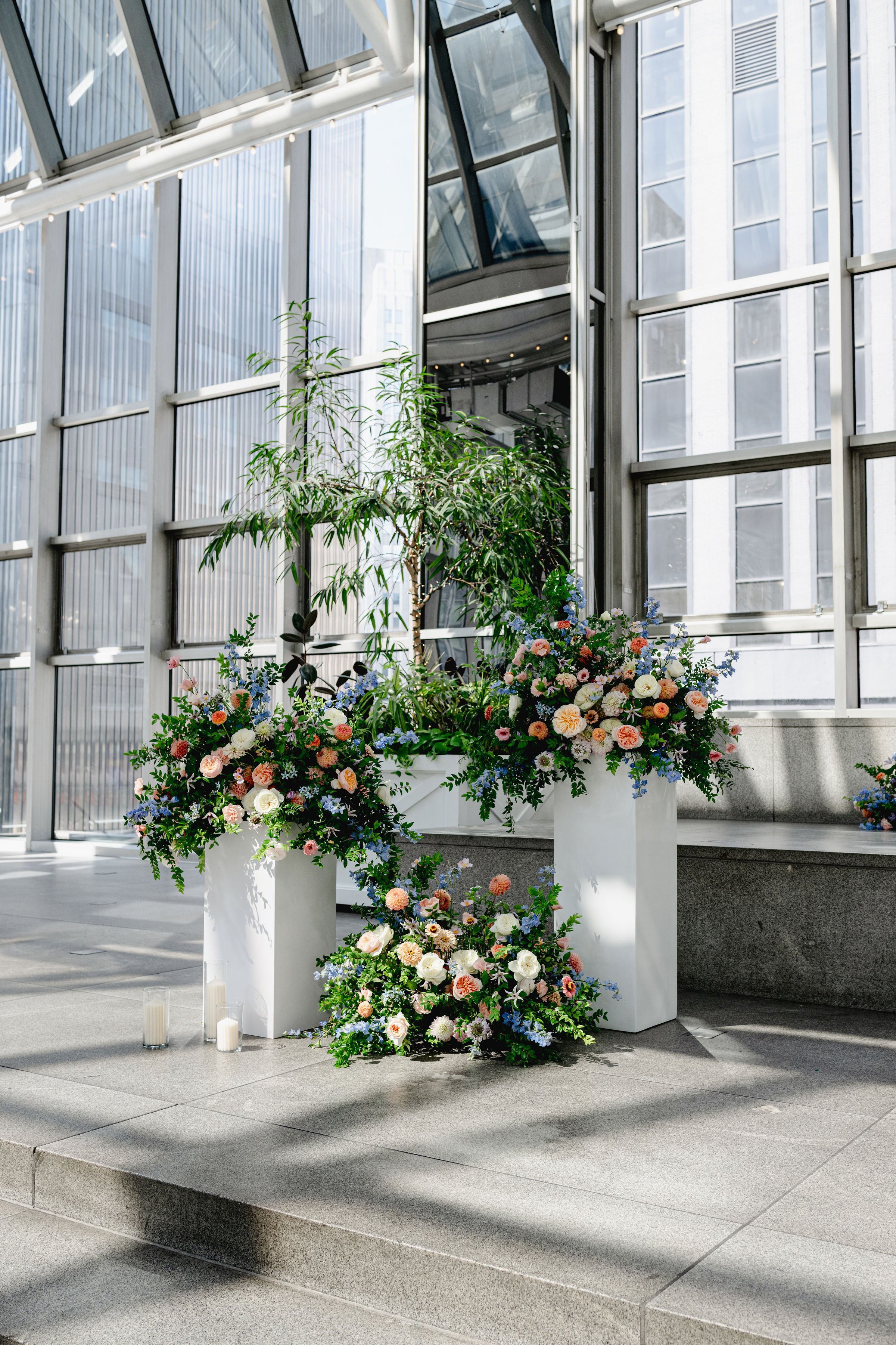 Indoor floral arrangement with colorful flowers and green foliage in white planters, set on a stone platform near a glass wall in a modern building.
