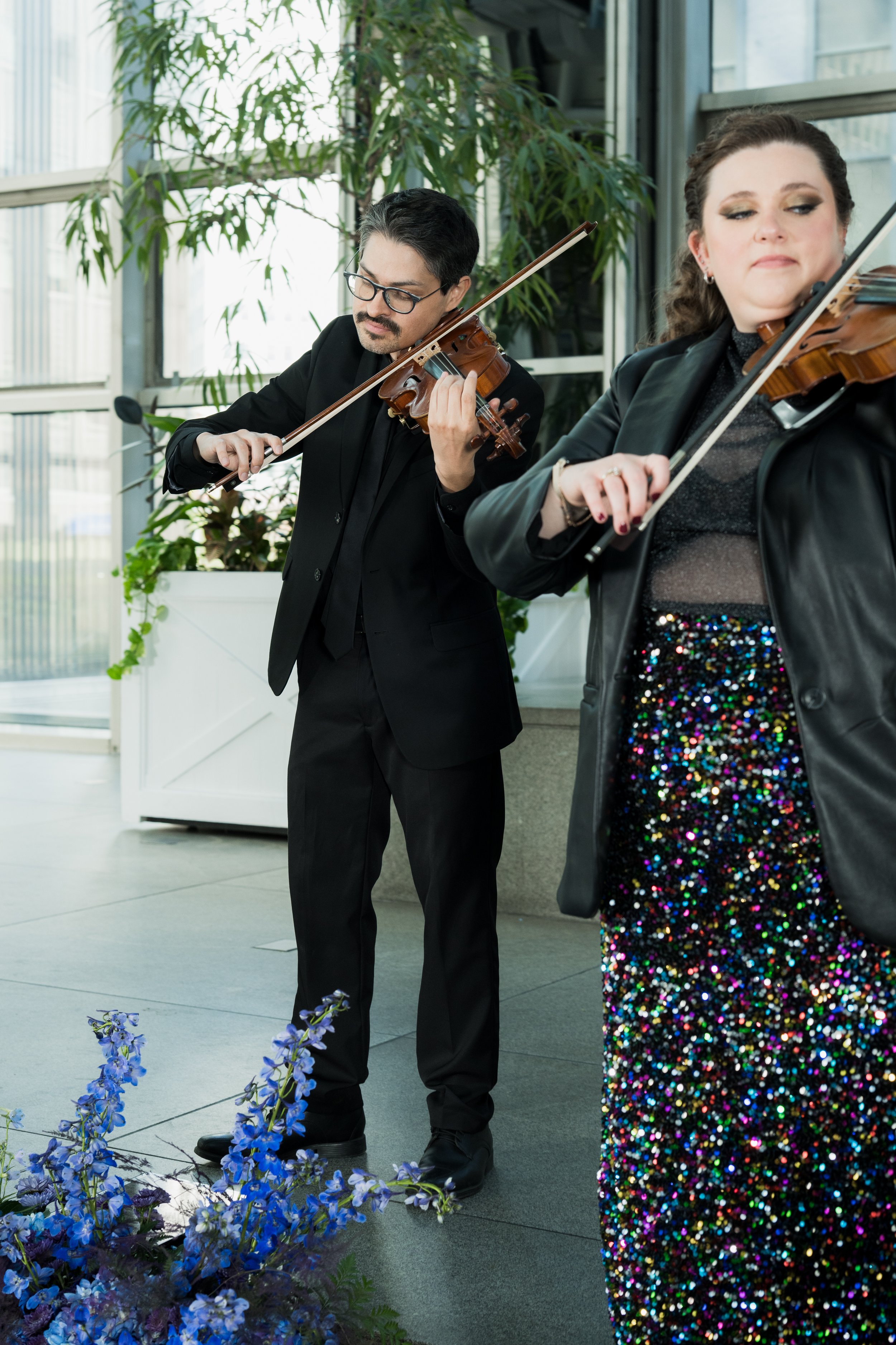 Musicians Hector Merchant and Nicole Lennartz playing violins indoors near large windows with greenery in the background.