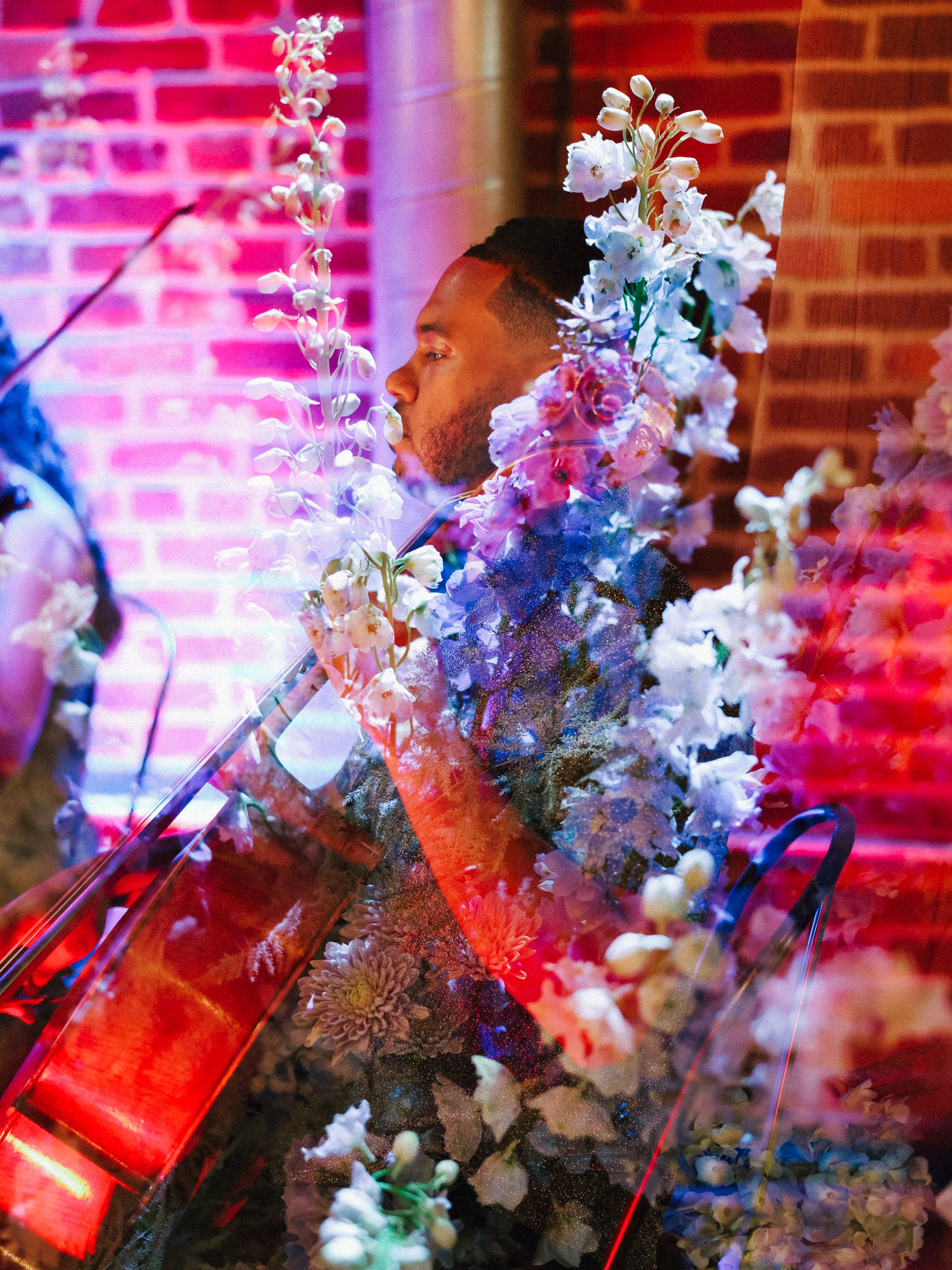 Cellist Ahmer'e Blackman sitting with his eyes closed, is surrounded by colorful flowers and reflections that create an abstract effect, with brick walls in the background.