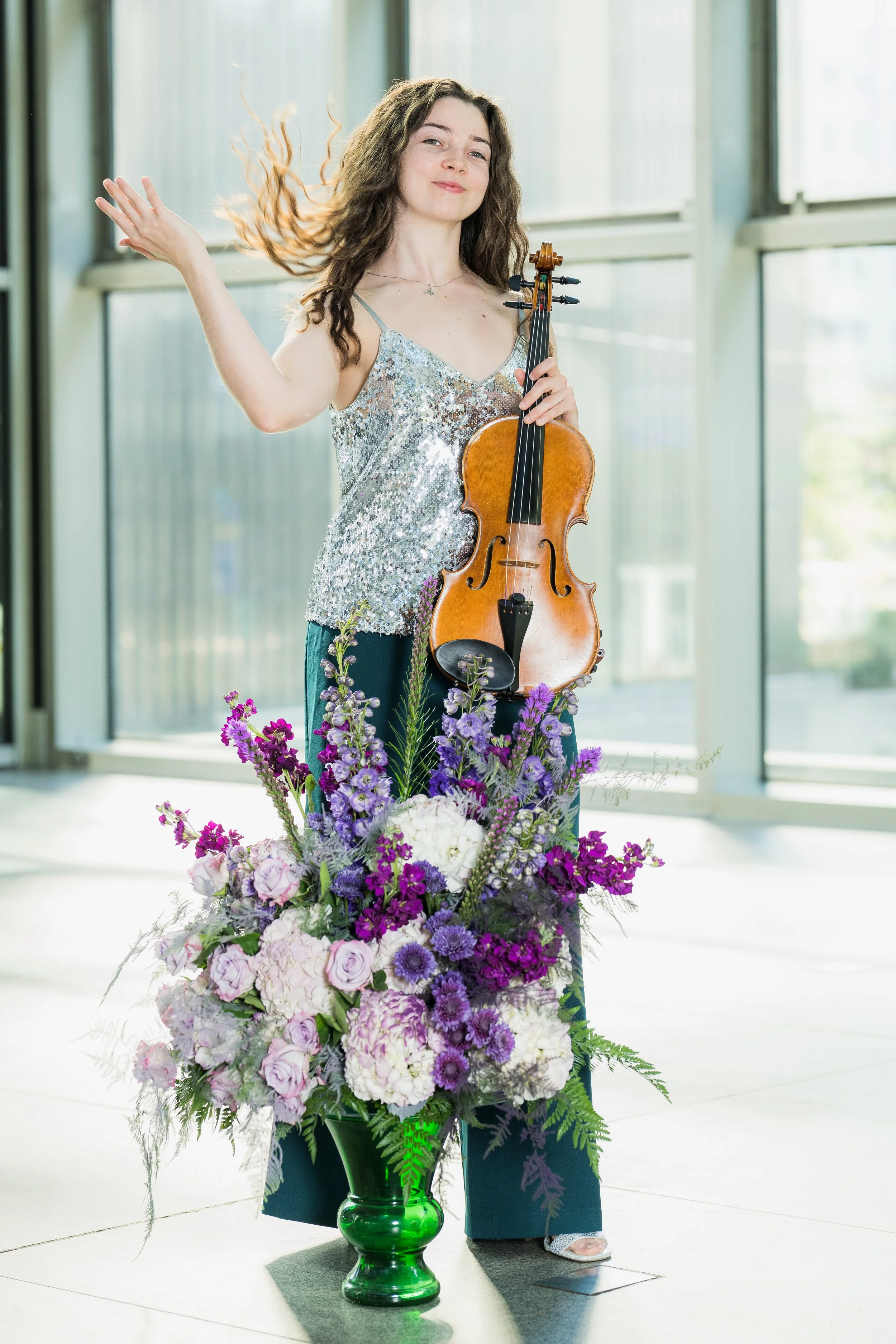 Erin Higgins holding a violin standing behind a large flower arrangement in an indoor space with large windows.